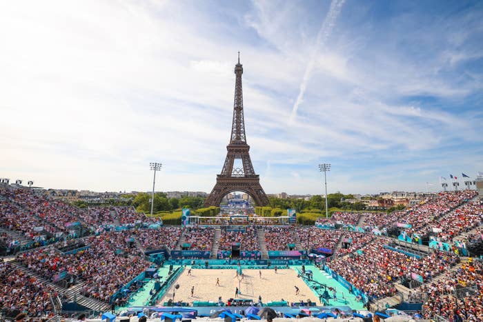 A packed stadium surrounds a beach volleyball court with the Eiffel Tower in the background, under a bright sky. Fans are watching the ongoing match