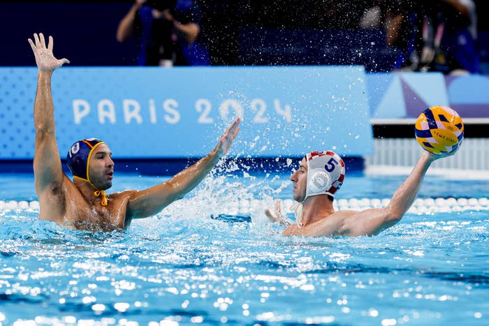 Two water polo players in action at the Paris 2024 event. One player (left side) raises arms defensively while the other (right side) prepares to throw the ball