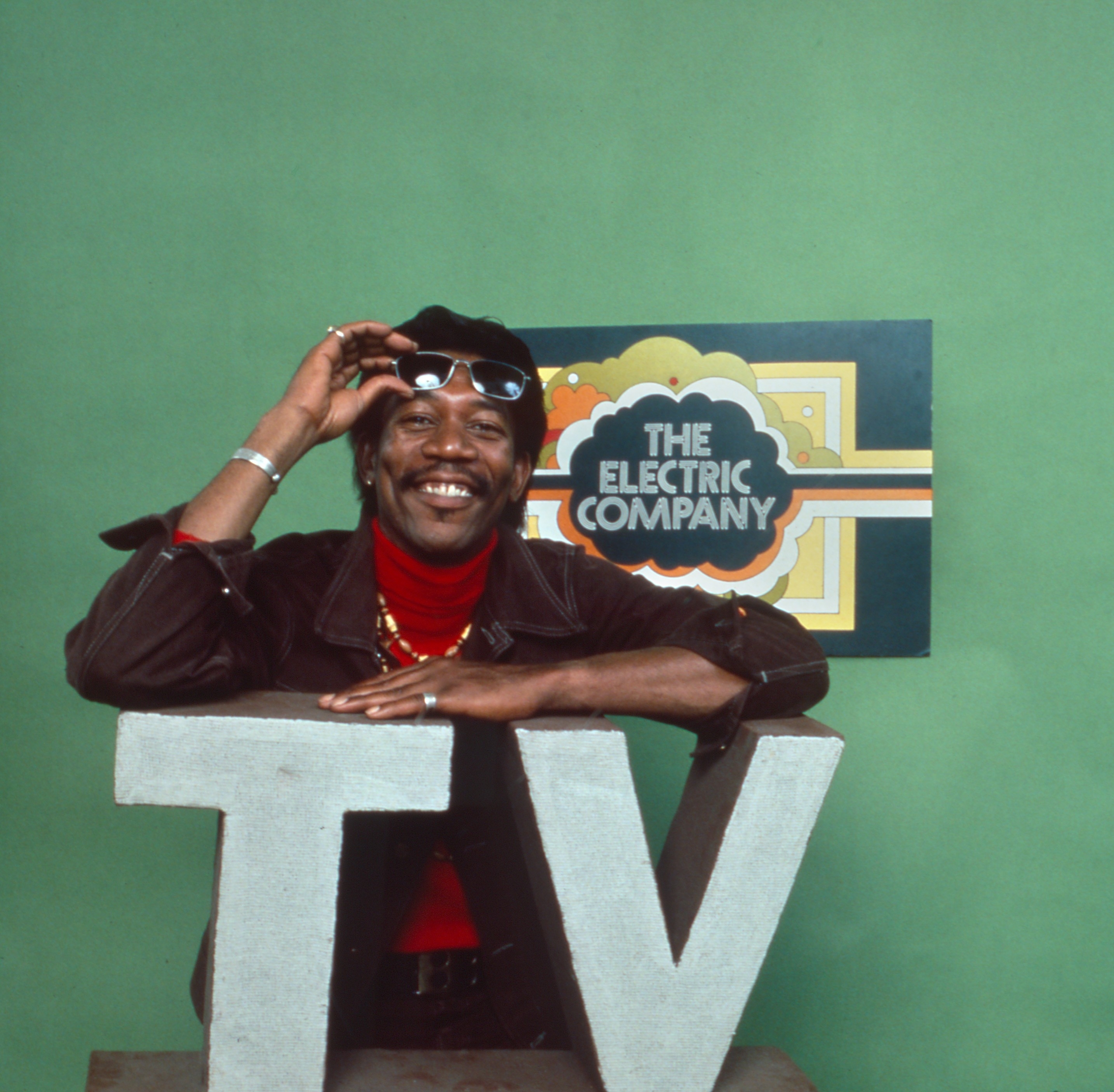 Morgan Freeman wearing a hat poses smiling with sunglasses perched on his head in front of a sign that reads "The Electric Company," leaning on large letters "TV"