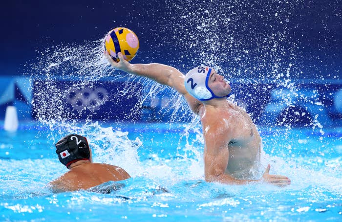 Two male water polo players in action; one jumps and throws the ball, while the other defends. The scene is from an Olympic water polo match, water splashing around