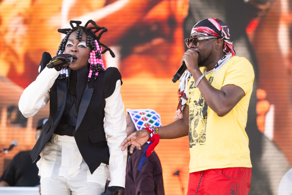 Lauryn Hill wearing a bold outfit with intricate braided hairstyle and Wyclef Jean in a bright t-shirt and bandana, performing on stage