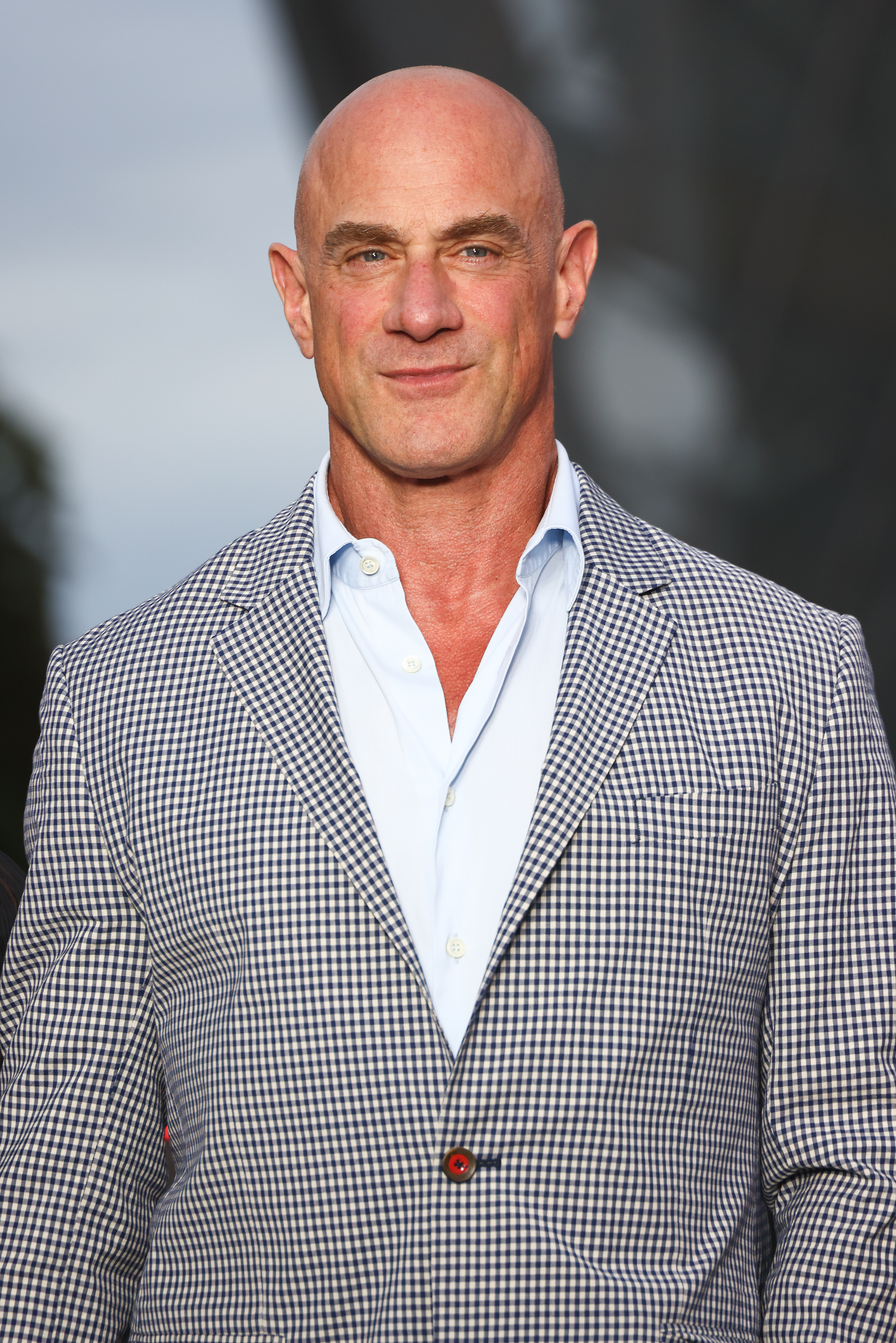 A bald man in a checkered blazer with a white shirt beneath, standing outdoors