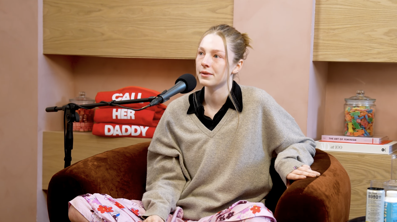 Hunter Schafer sits in an armchair during a podcast interview, wearing a casual sweater and floral pants