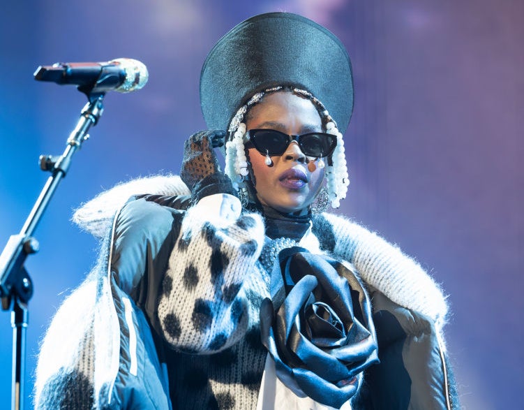Lauryn Hill in a unique outfit with a large hat, sunglasses, and a patterned jacket, performing on stage holding a microphone