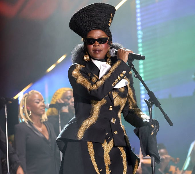 Lauryn Hill performs on stage wearing a patterned black and gold suit, a tall black hat, and sunglasses. Backup singers in similar black attire are in the background