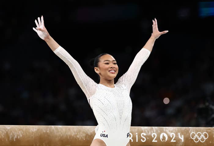 Gymnast Suni Lee celebrates with arms raised after completing a routine at the Paris 2024 Olympics. She is wearing a white leotard with USA written on it
