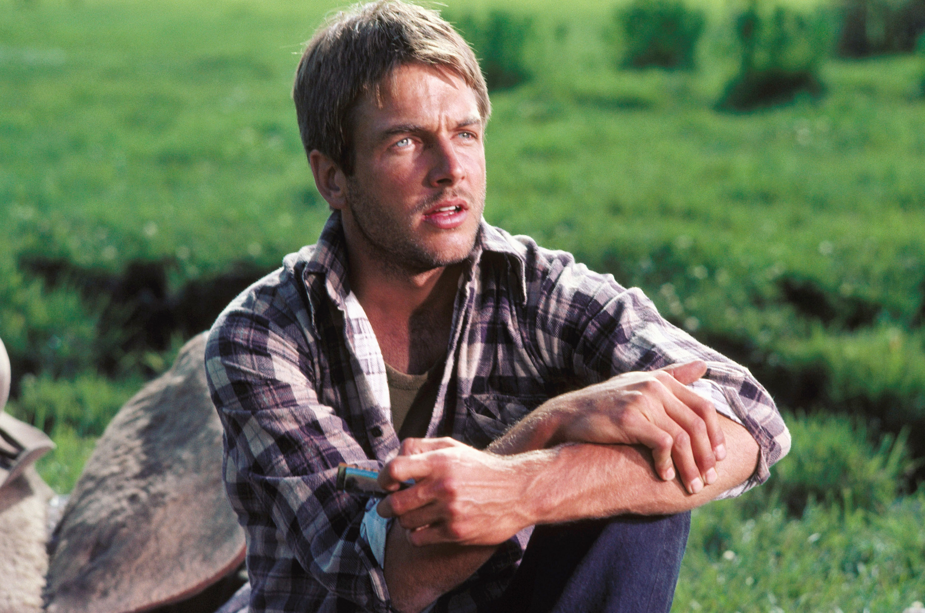 Mark Harmon, wearing a plaid shirt and jeans, sits outdoors on grassy terrain, looking thoughtful.