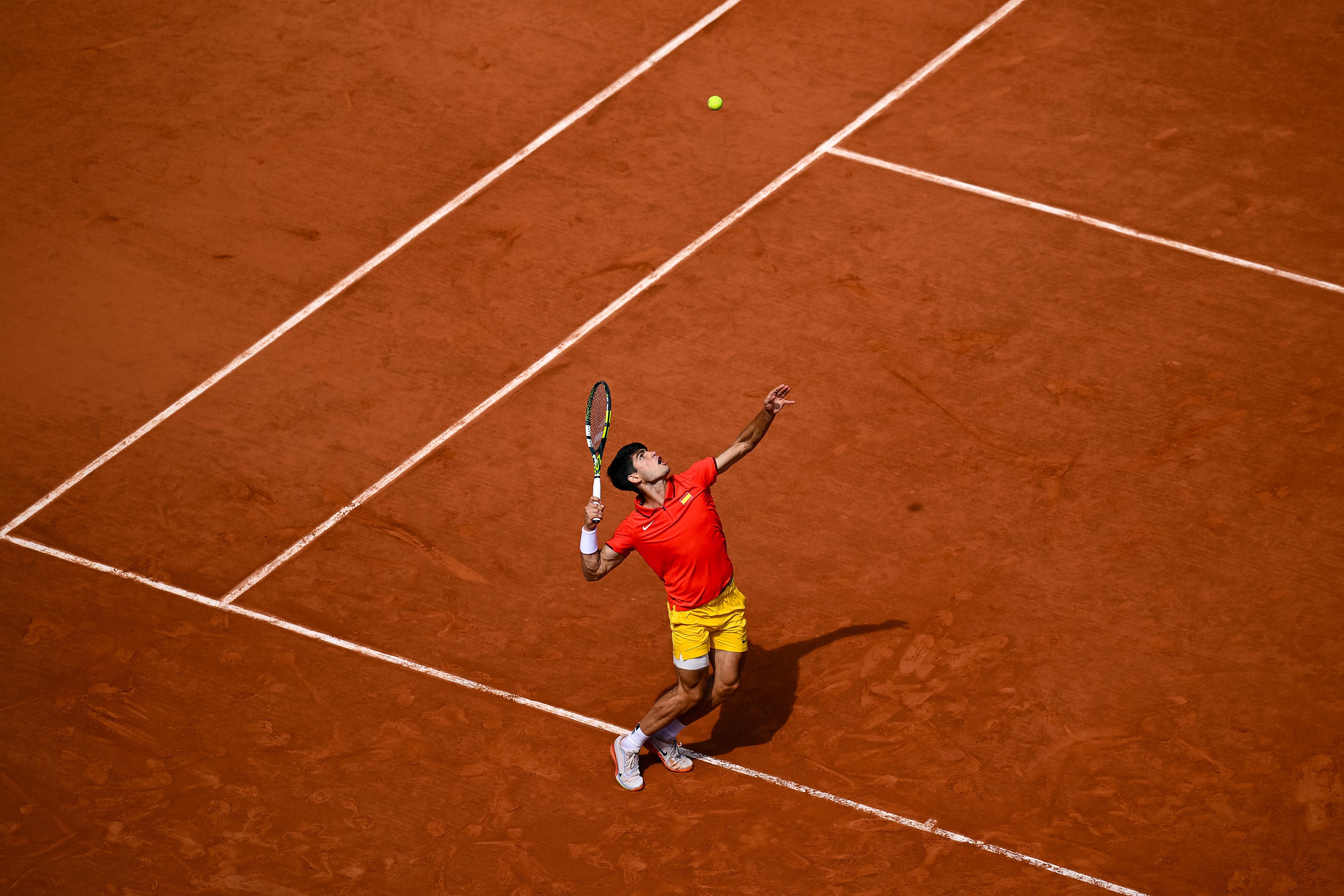Tennis player Novak Djokovic in action, wearing a red shirt and yellow shorts, preparing to serve a tennis ball on a clay court