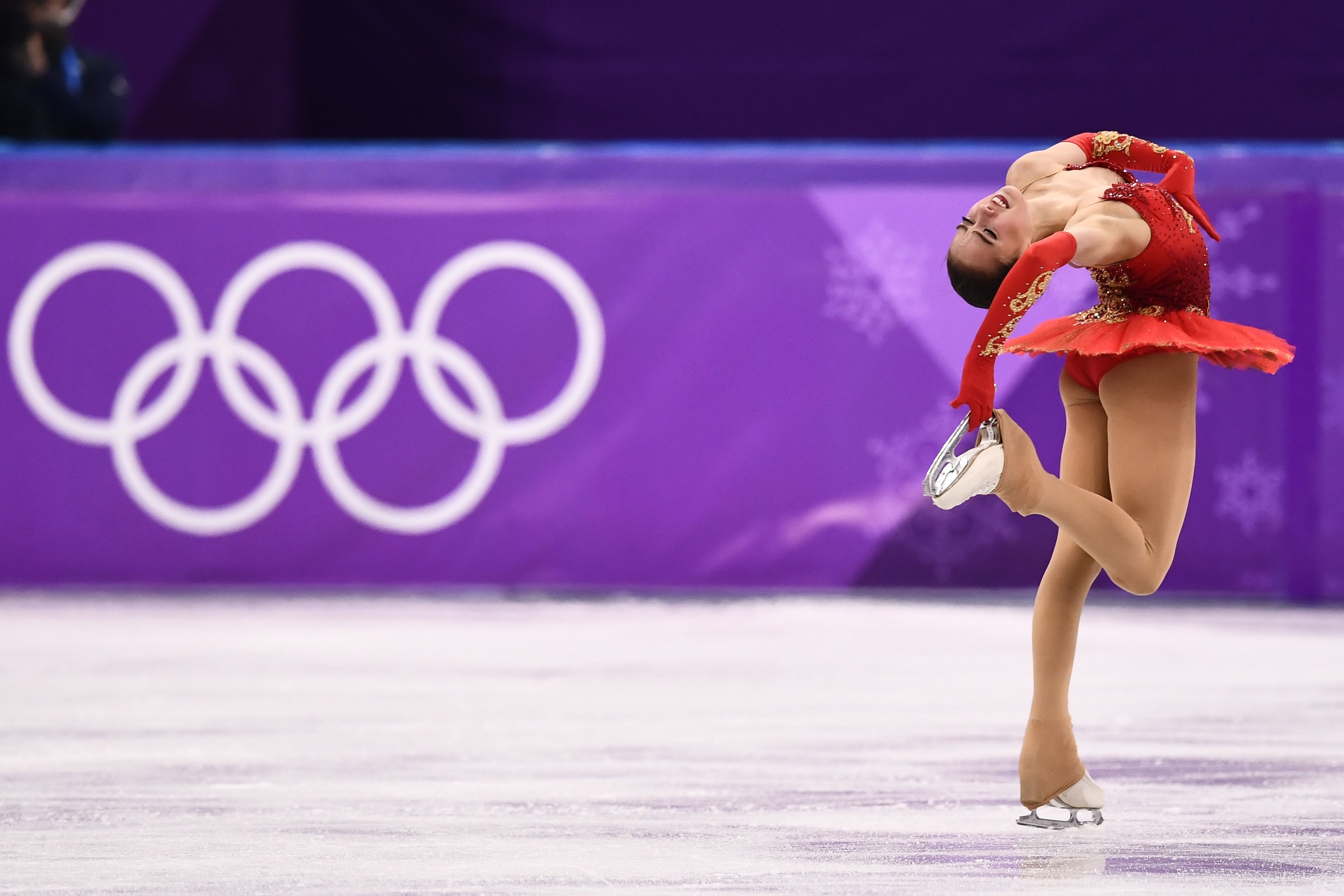 Figure skater in mid-performance wearing an elegant dress, with the Olympic rings in the background