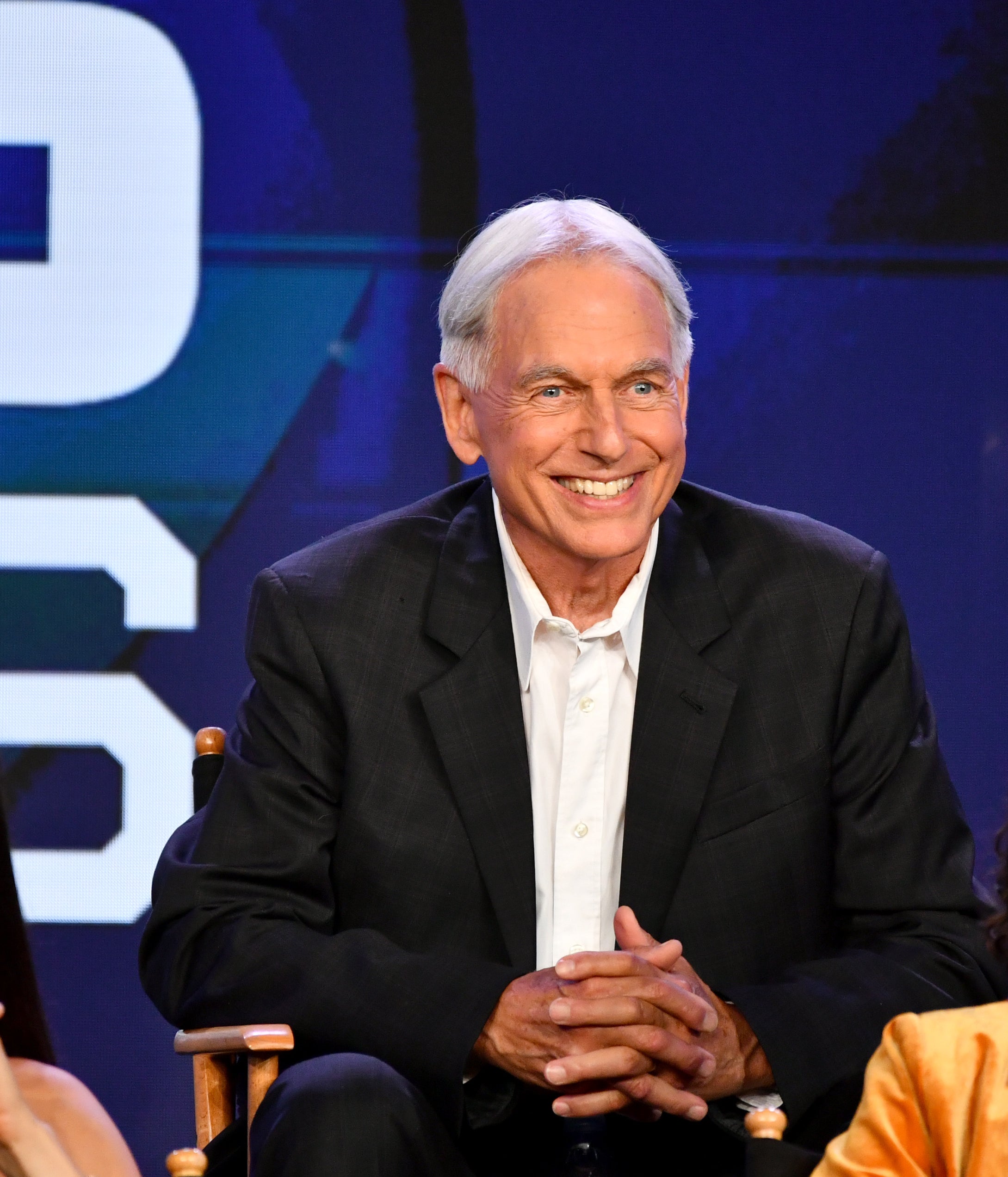 Mark Harmon in a black suit sits between two women, one in a sleeveless top and the other in a yellow blazer, at an event panel