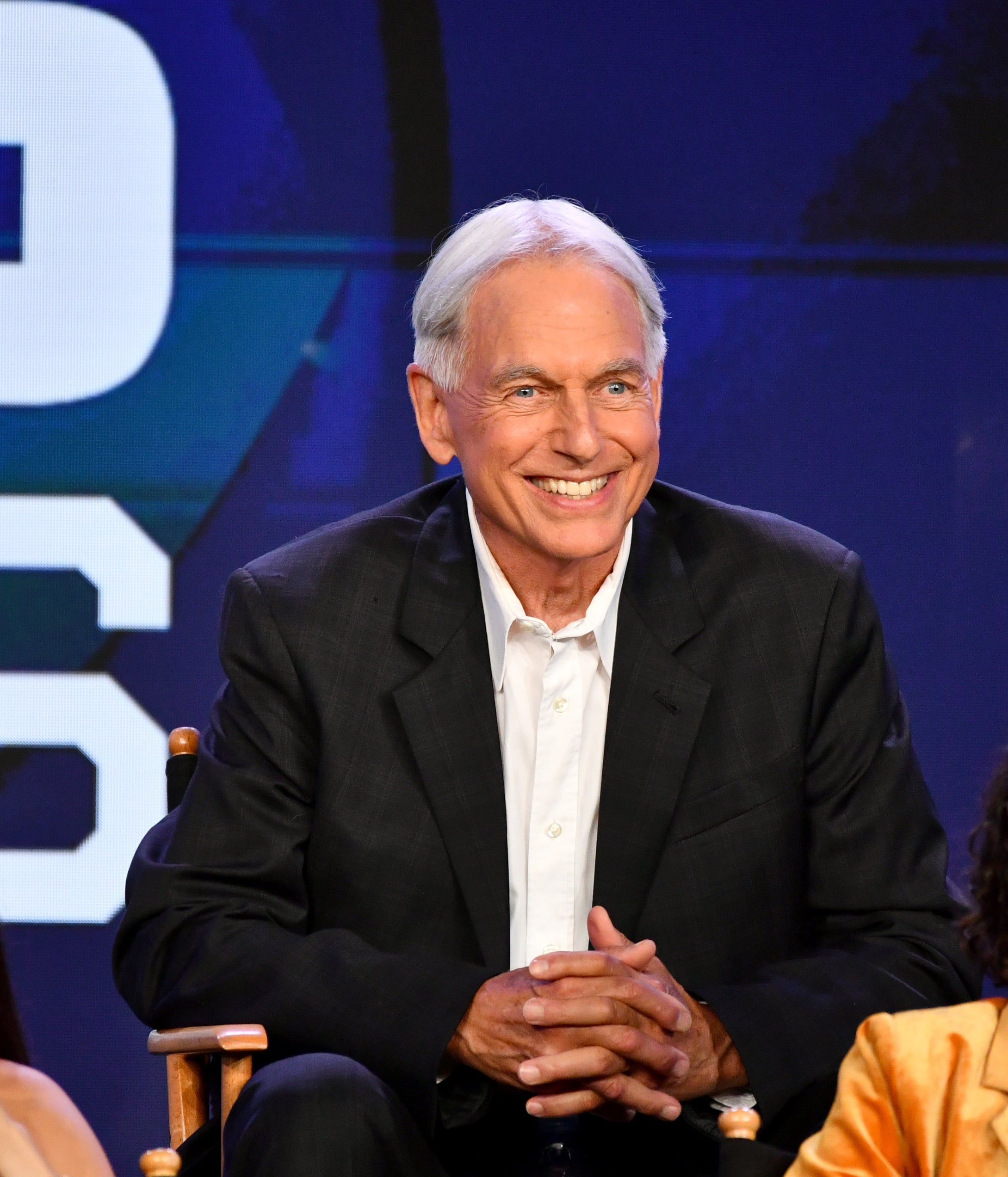 Mark Harmon in a black suit sits between two women, one in a sleeveless top and the other in a yellow blazer, at an event panel
