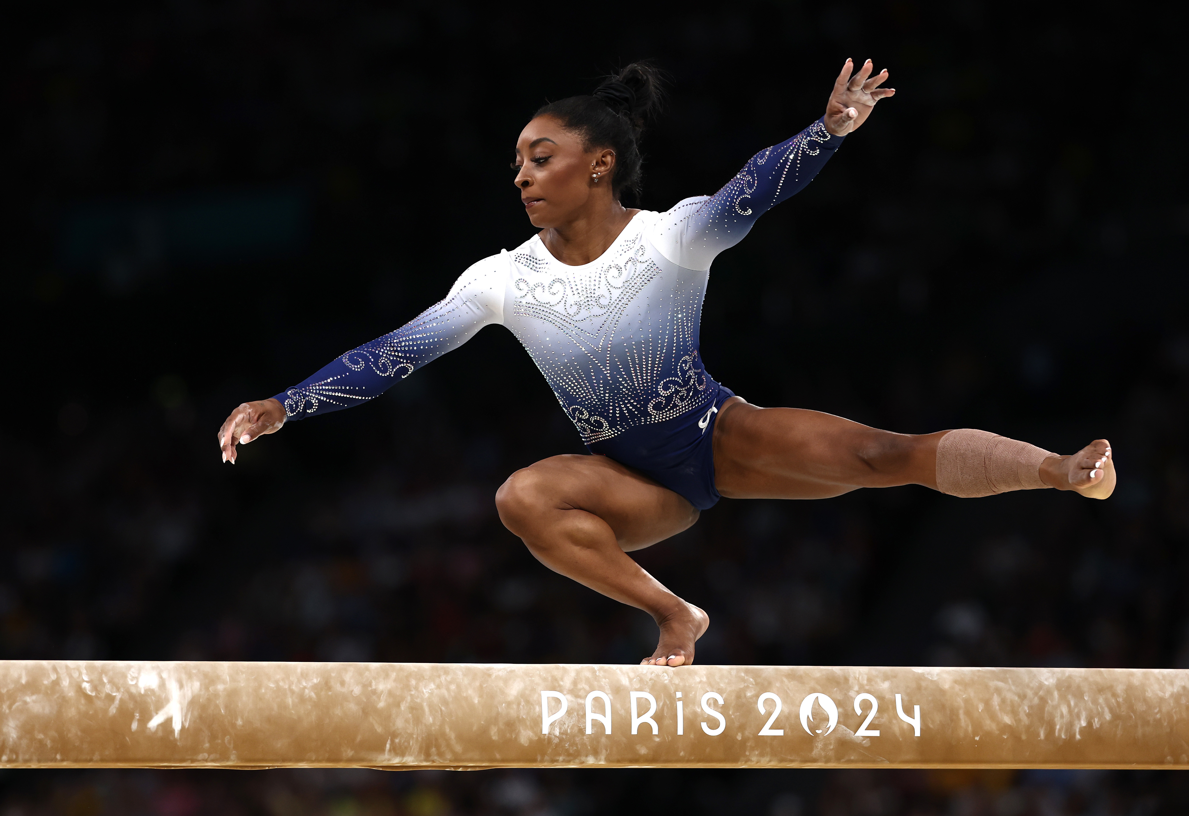 Simone Biles performs on the balance beam at the 2024 Paris Olympics, wearing a leotard with a gradient design and her right leg bandaged