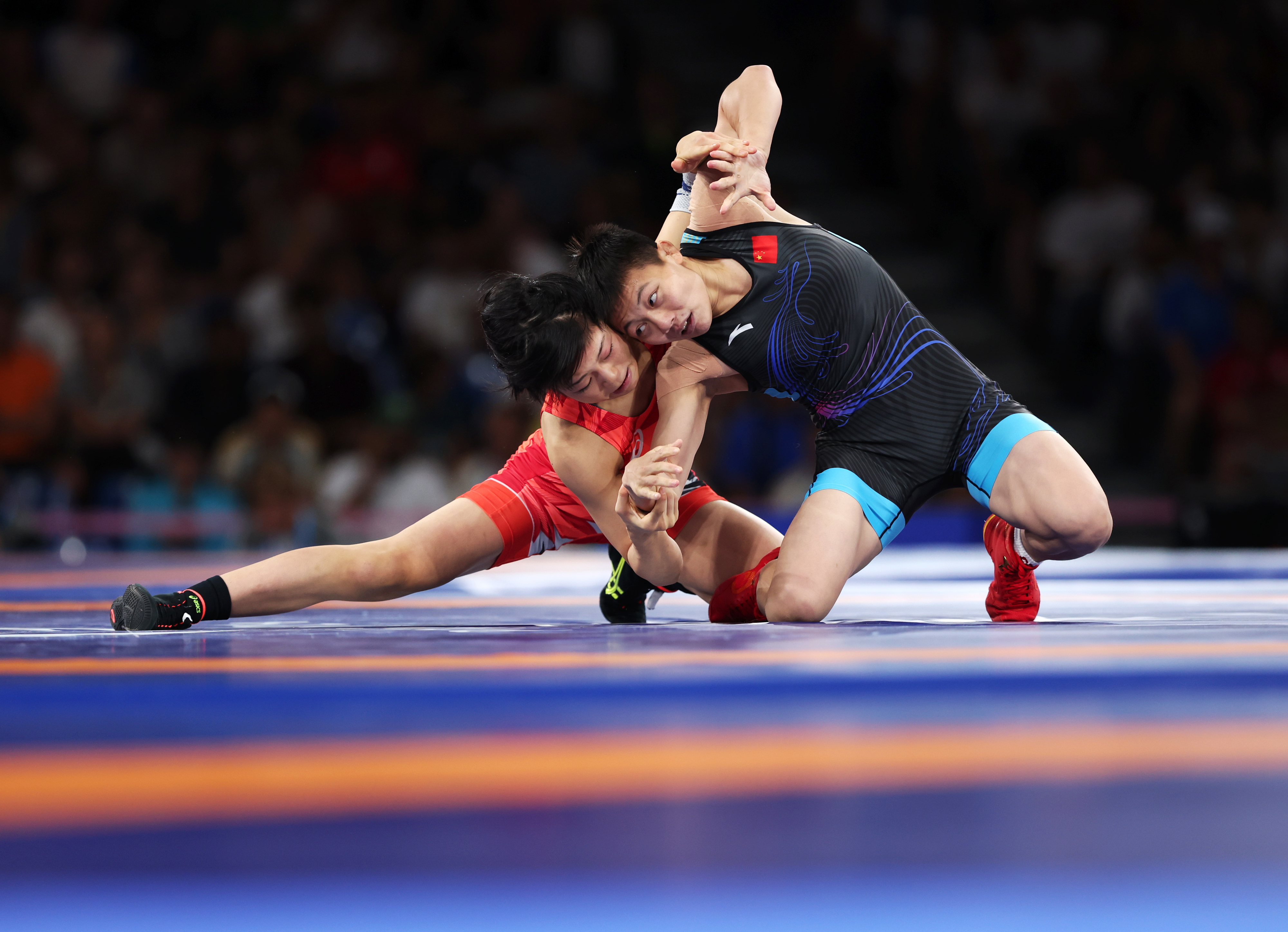 Two female wrestlers in action on a wrestling mat during a match. One is in a red outfit, and the other in a black and blue outfit, both grappling intensely
