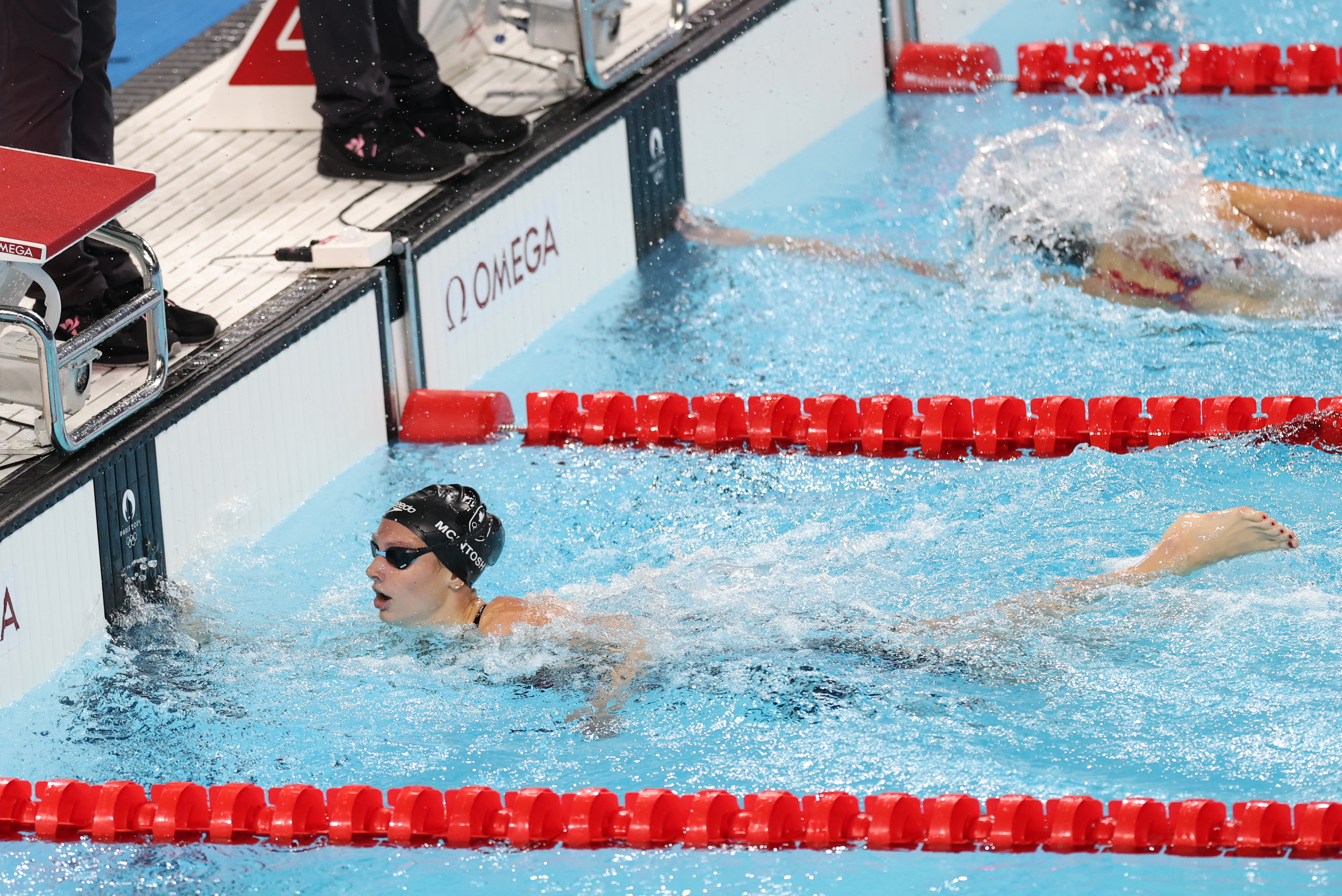 Swimmer touching the wall in a swimming race, signifying the end of a lap. Another swimmer is seen behind, creating splashes in the pool