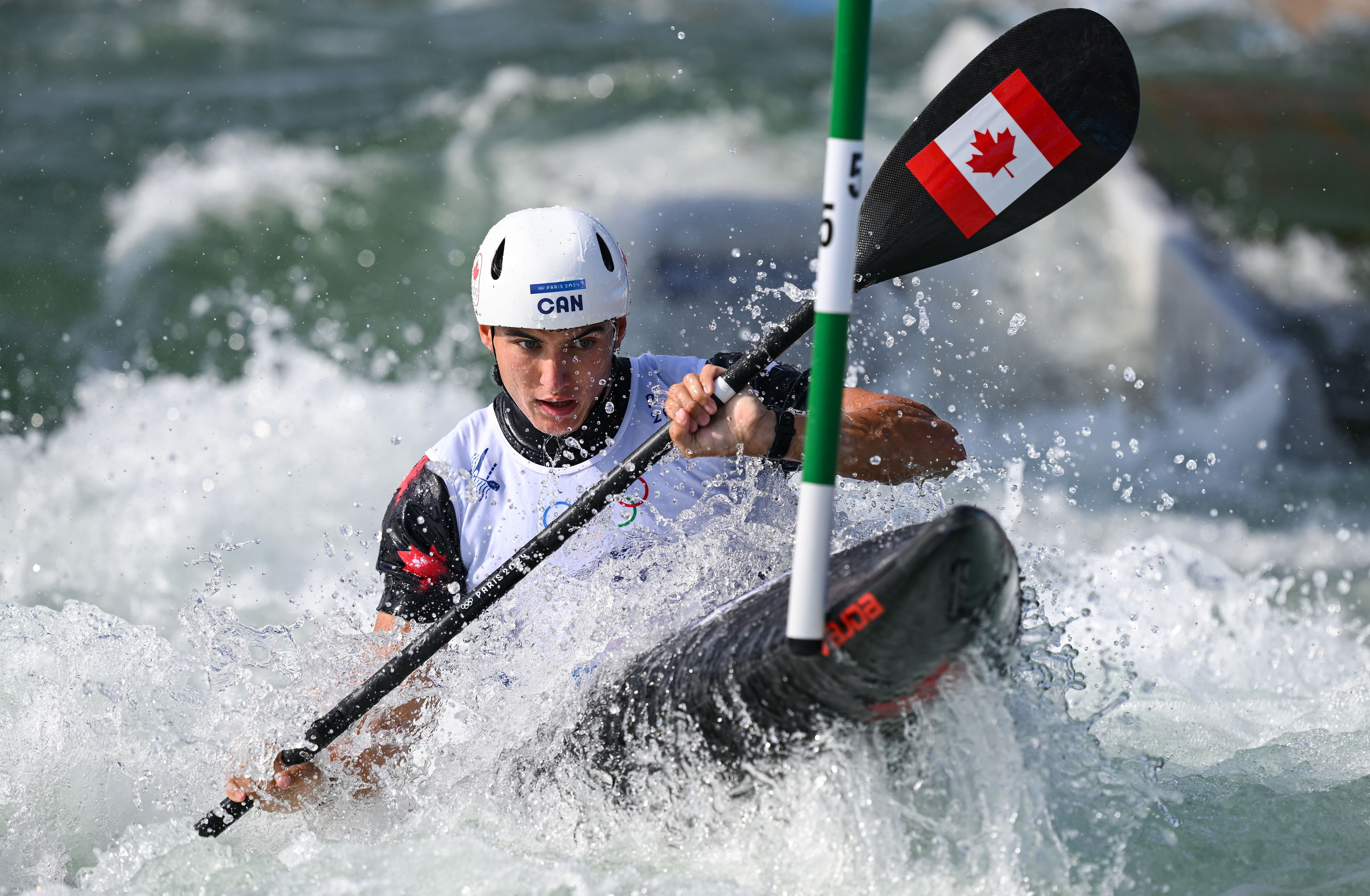 A person in white helmet and kayak gear is navigating through whitewater rapids in a kayak with a Canadian flag on the paddle