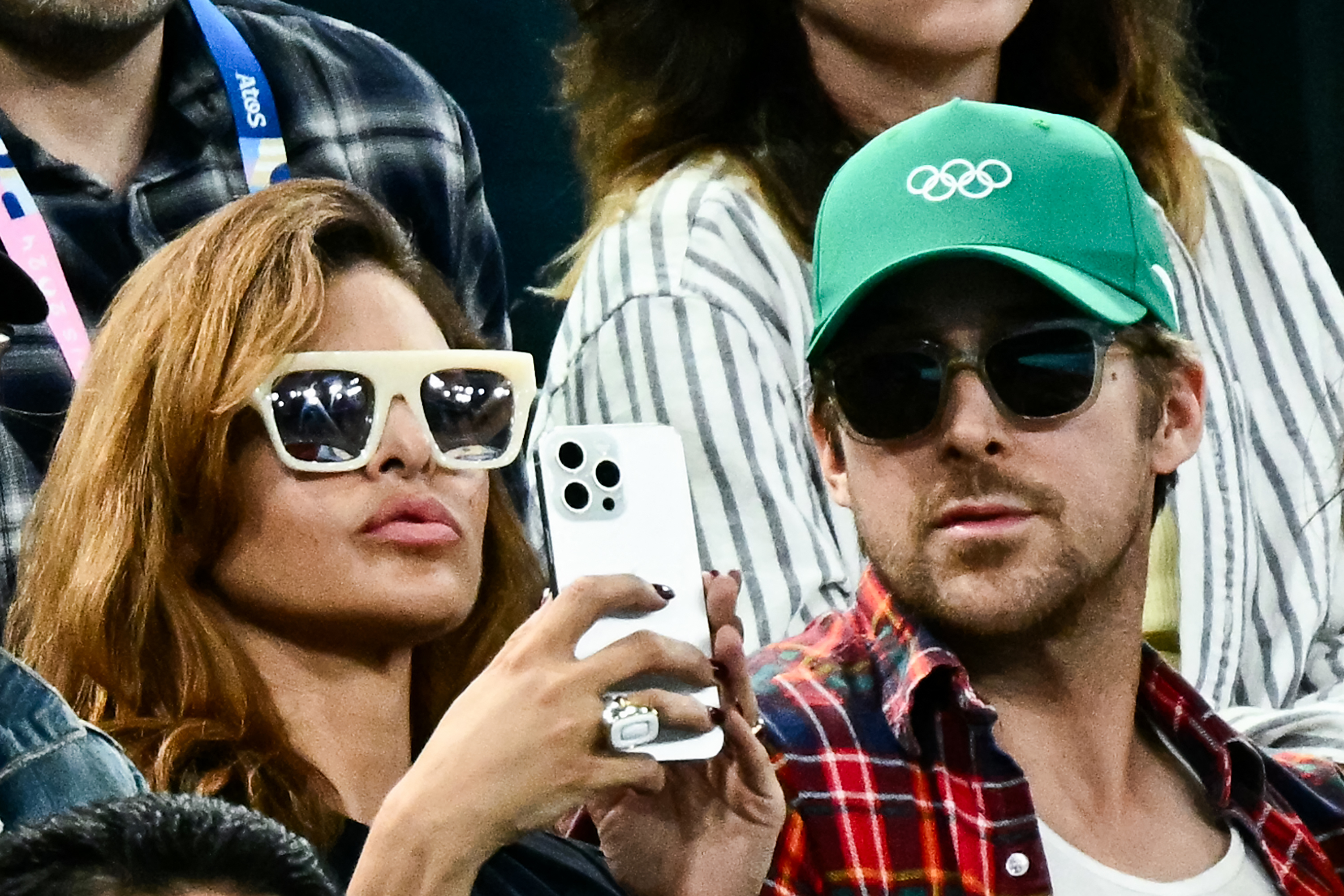 Eva Mendes and Ryan Gosling are at Olympics; Eva wears sunglasses and holds a phone, Ryan wears a hat and sunglasses, they are seated among others