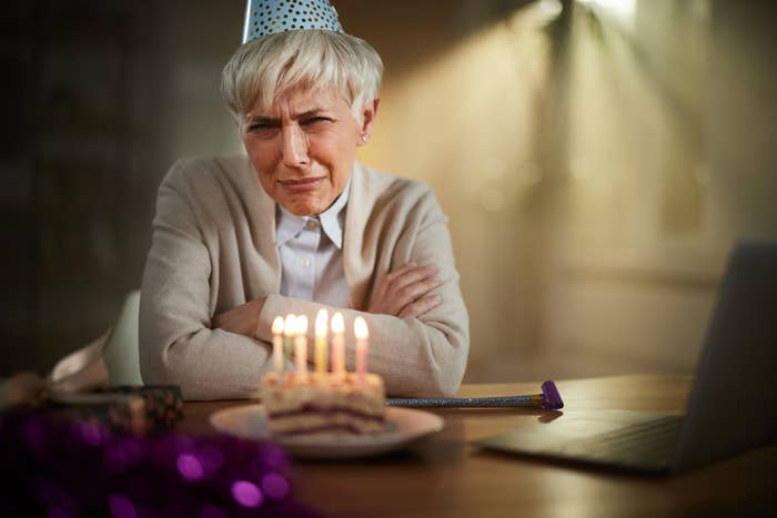 An older person with short, white hair is sitting at a table wearing a party hat, looking unimpressed at a lit birthday cake with candles in front of them