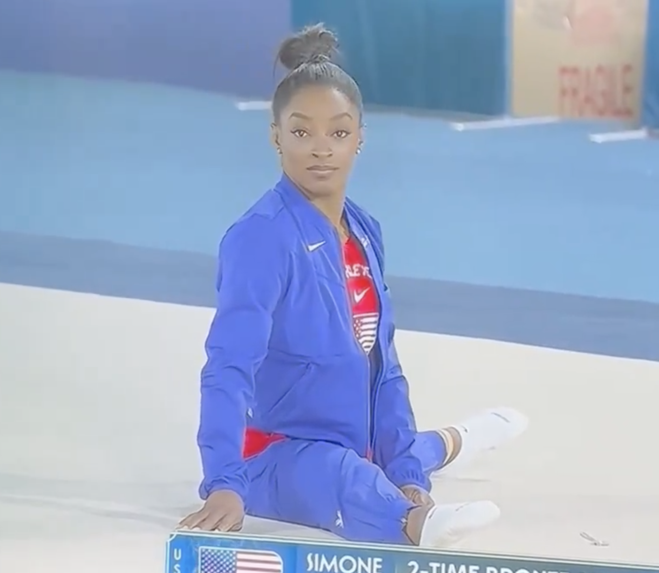 Simone Biles sitting on a gymnastics mat in a blue tracksuit, looking towards the camera. She is at a sporting event, indicated by the sign next to her