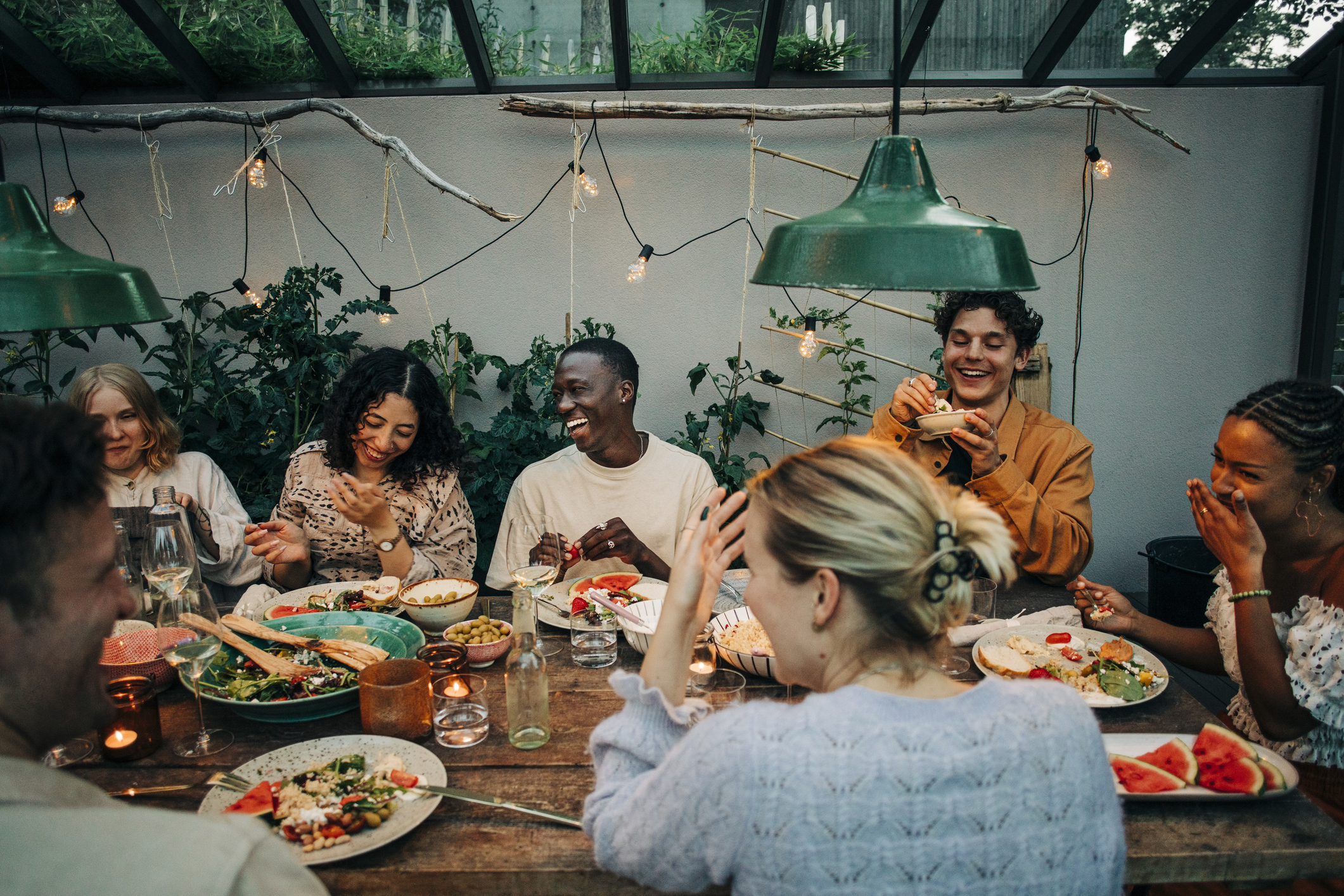 Group of people having a joyful meal together at an outdoor table under hanging lights and surrounded by plants