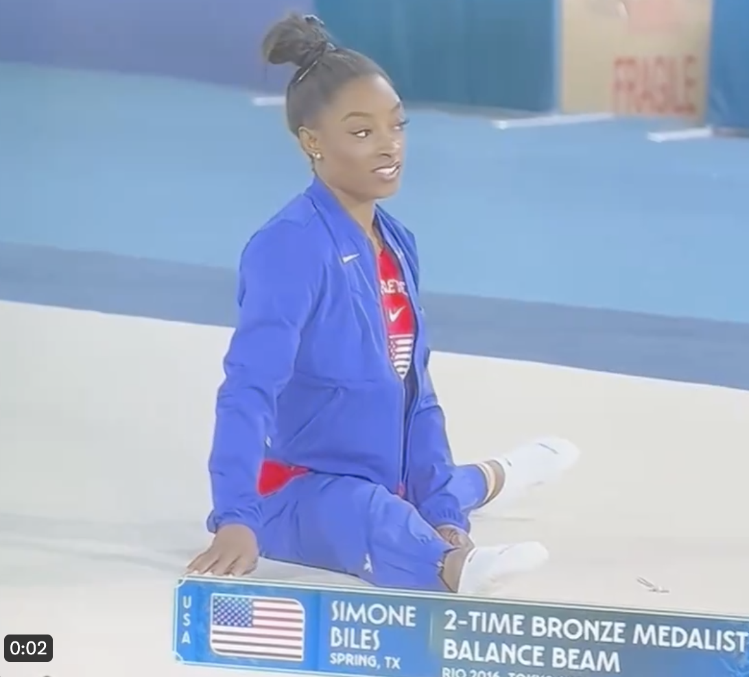 Simone Biles, in athletic wear, sits by a sign noting her as a 2-time bronze medalist in the balance beam