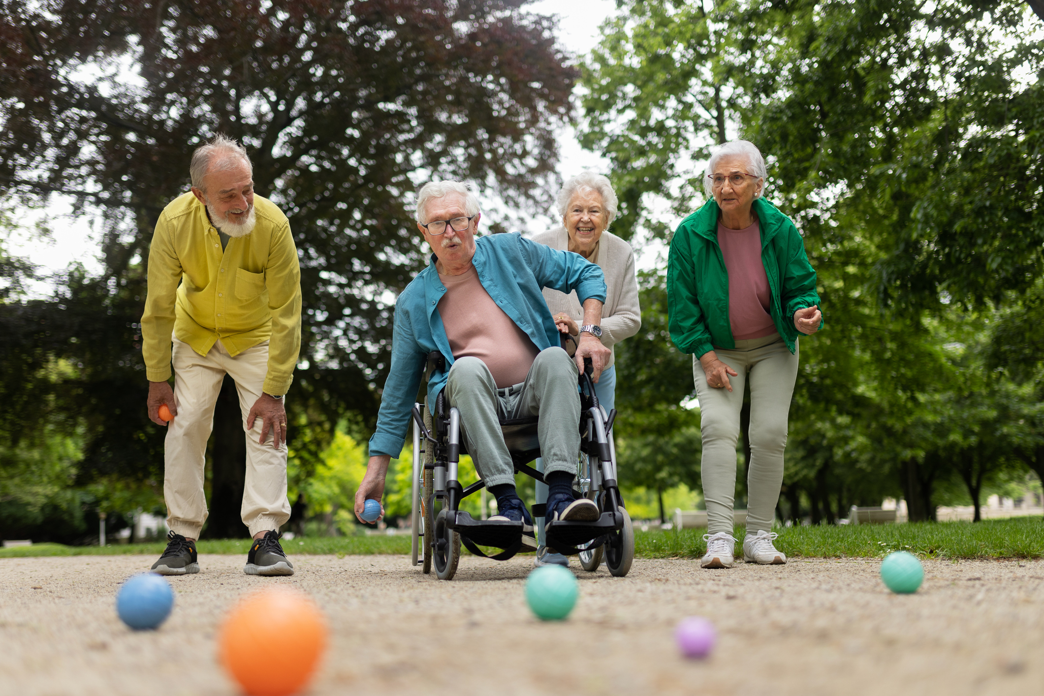 An elderly man in a wheelchair playing bocce with three elderly friends in a park. All appear joyful and engaged in the game