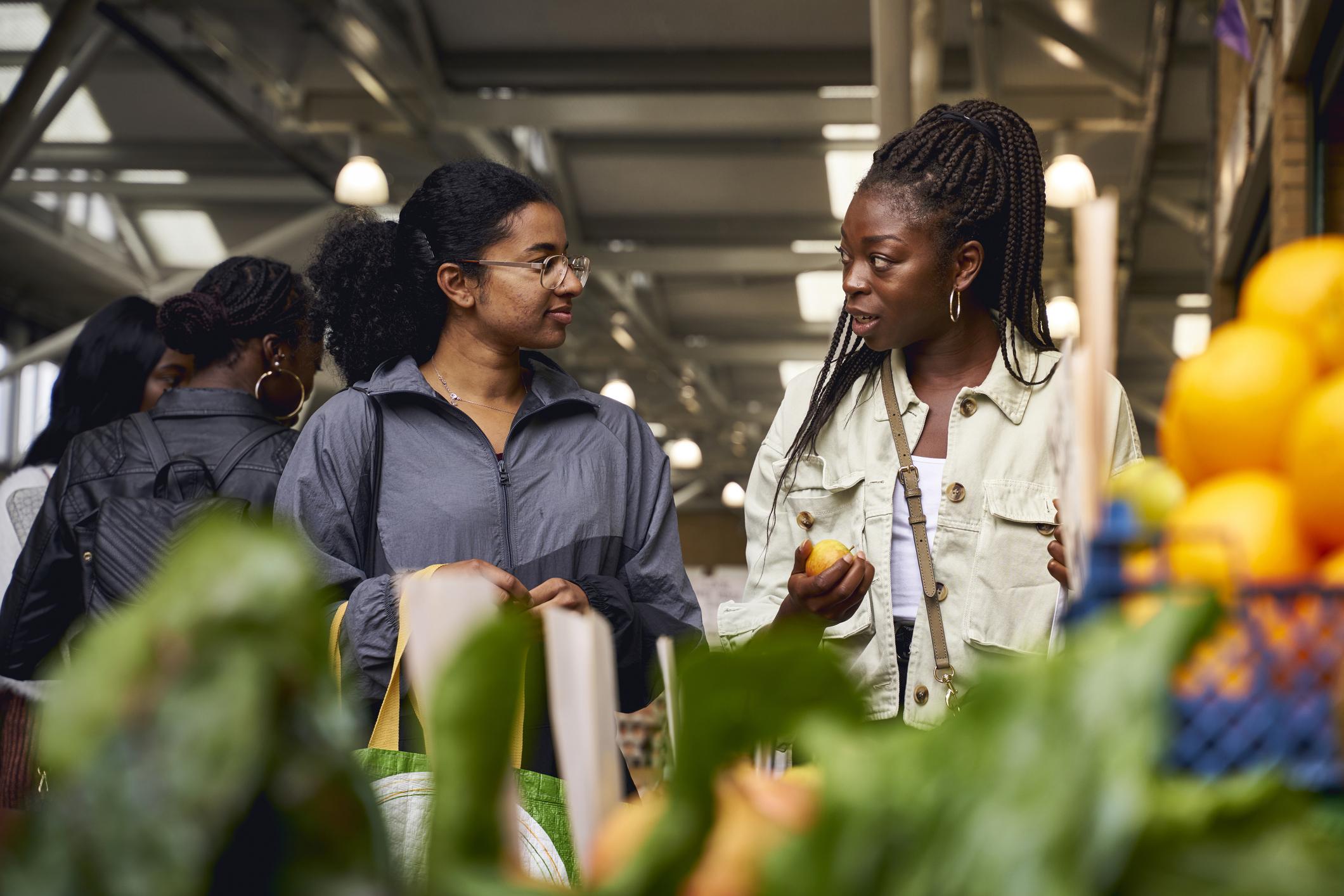 Two women engage in conversation while grocery shopping at a market, one holding an orange