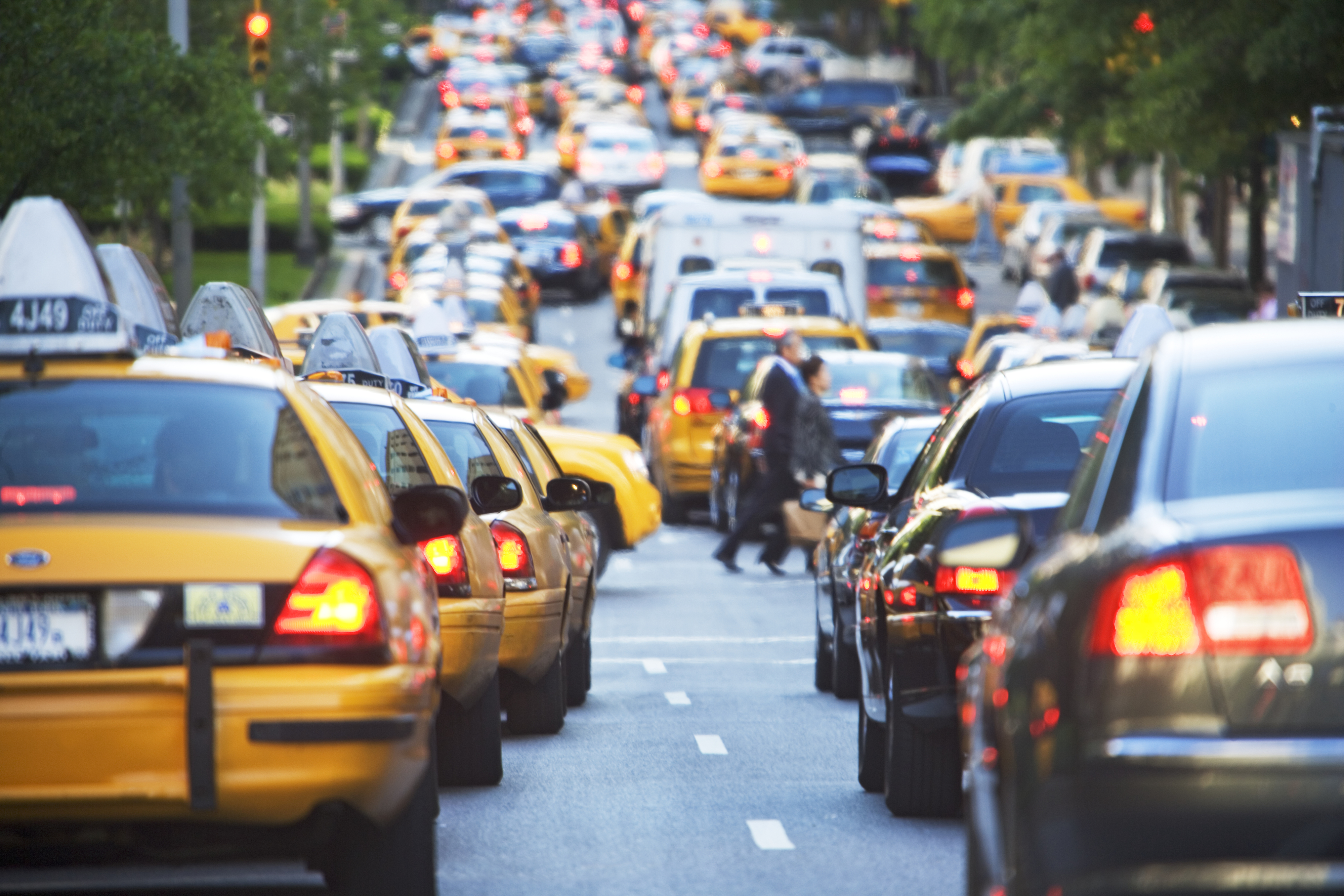 A busy city street filled with yellow taxis and other cars. Pedestrians cross between the vehicles amidst the traffic congestion