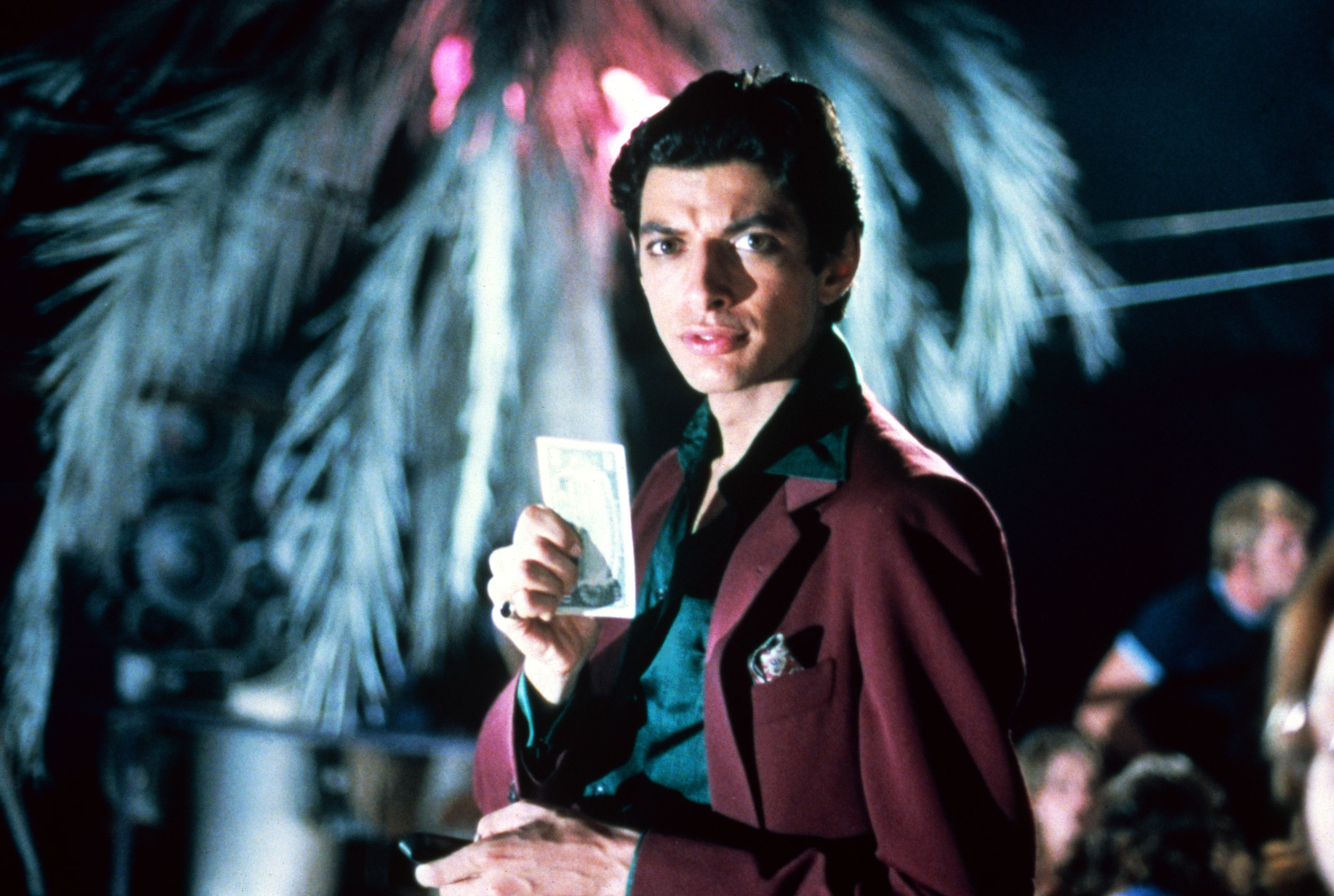 Young Jeff Goldblum, in a suit, holds a dollar bill, looking at the camera. The background has a palm tree and blurred figures