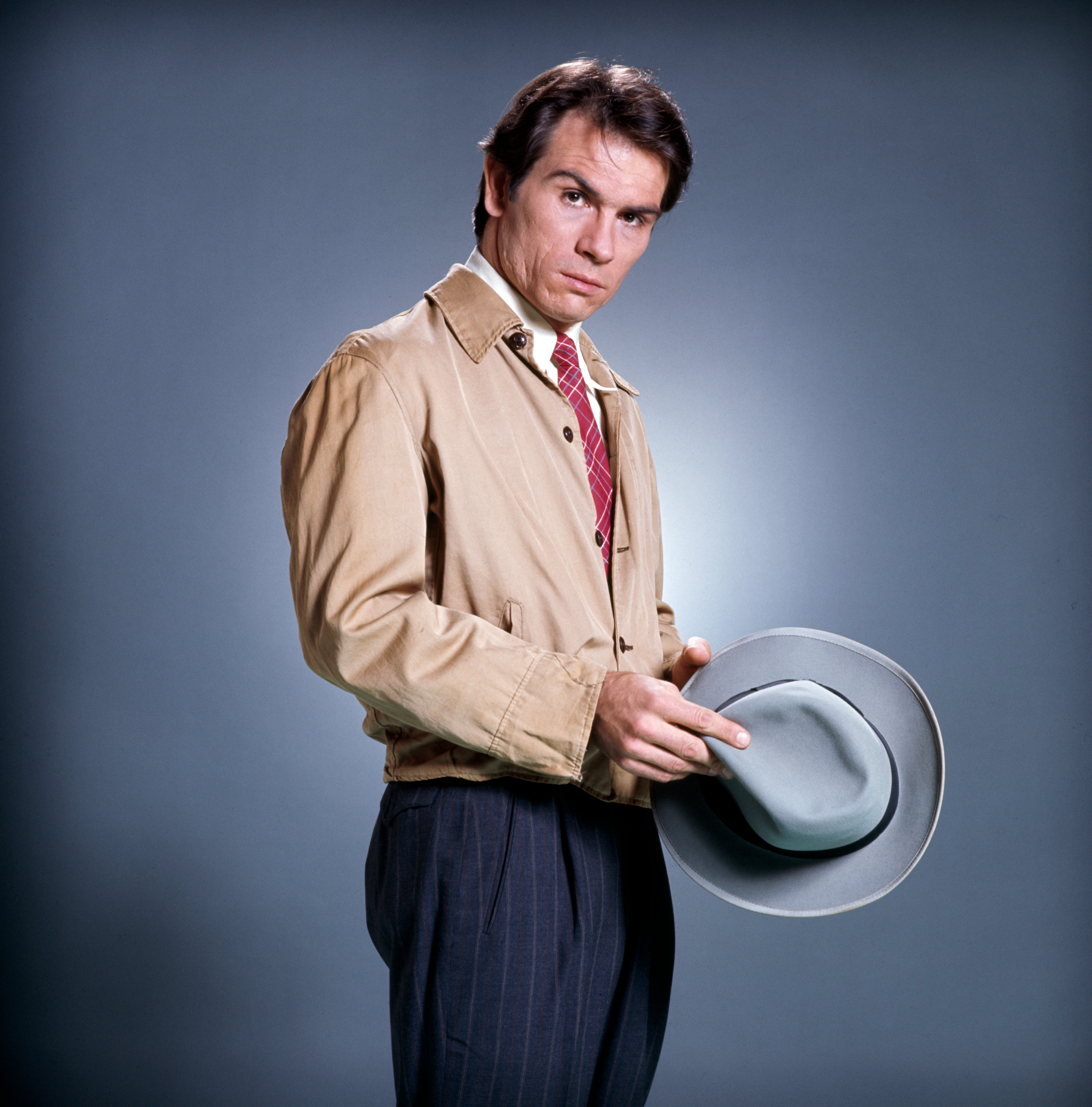 Sam Neill in a tan jacket, tie, and striped pants, holding a gray hat against a plain backdrop