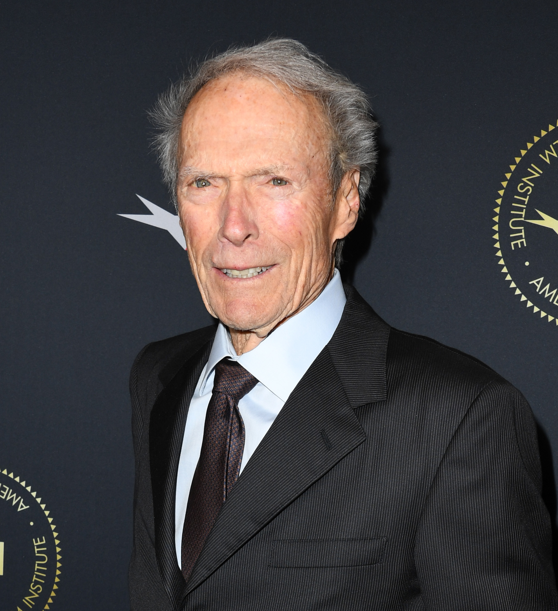 Clint Eastwood at an event, wearing a dark suit with a white shirt and a dark tie, standing in front of a backdrop with gold emblems
