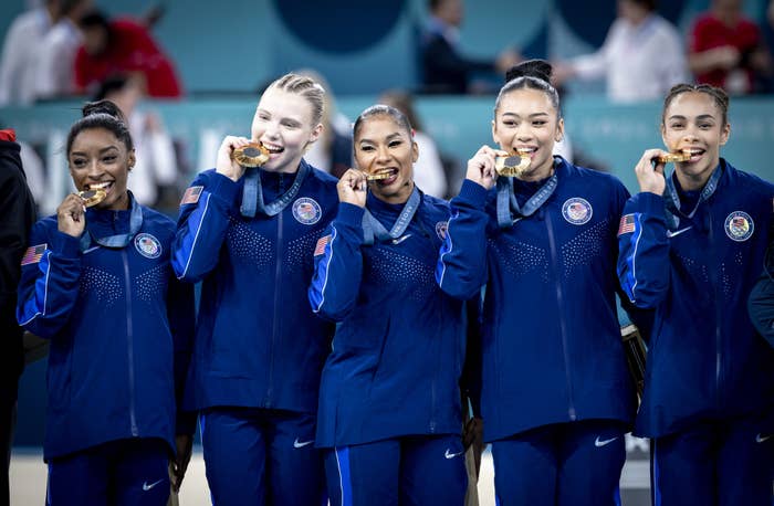 Simone Biles, Grace McCallum, Sunisa Lee, Jordan Chiles, and Jade Carey pose with gold medals at a gymnastics event, all wearing matching team tracksuits
