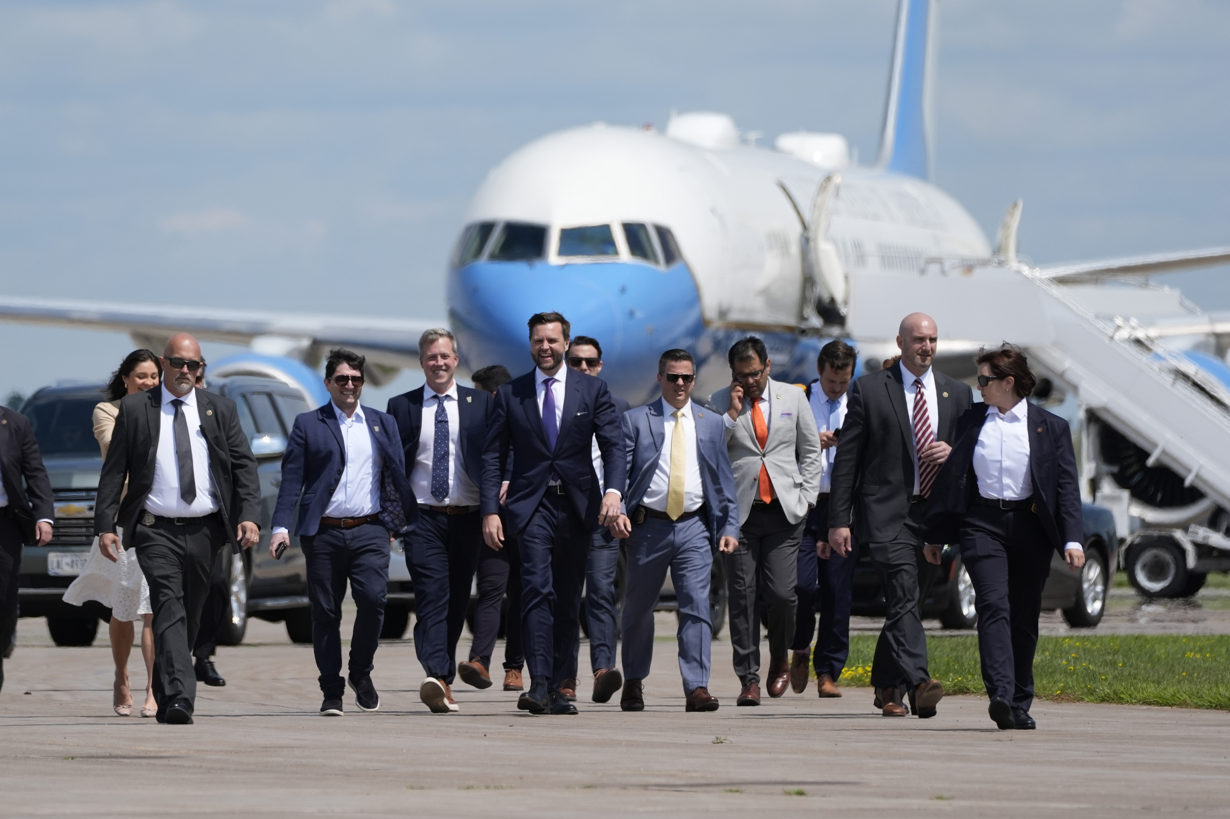 Nine people, all in business attire including suits and ties, walk on an airport tarmac with a large airplane in the background. Names undisclosed