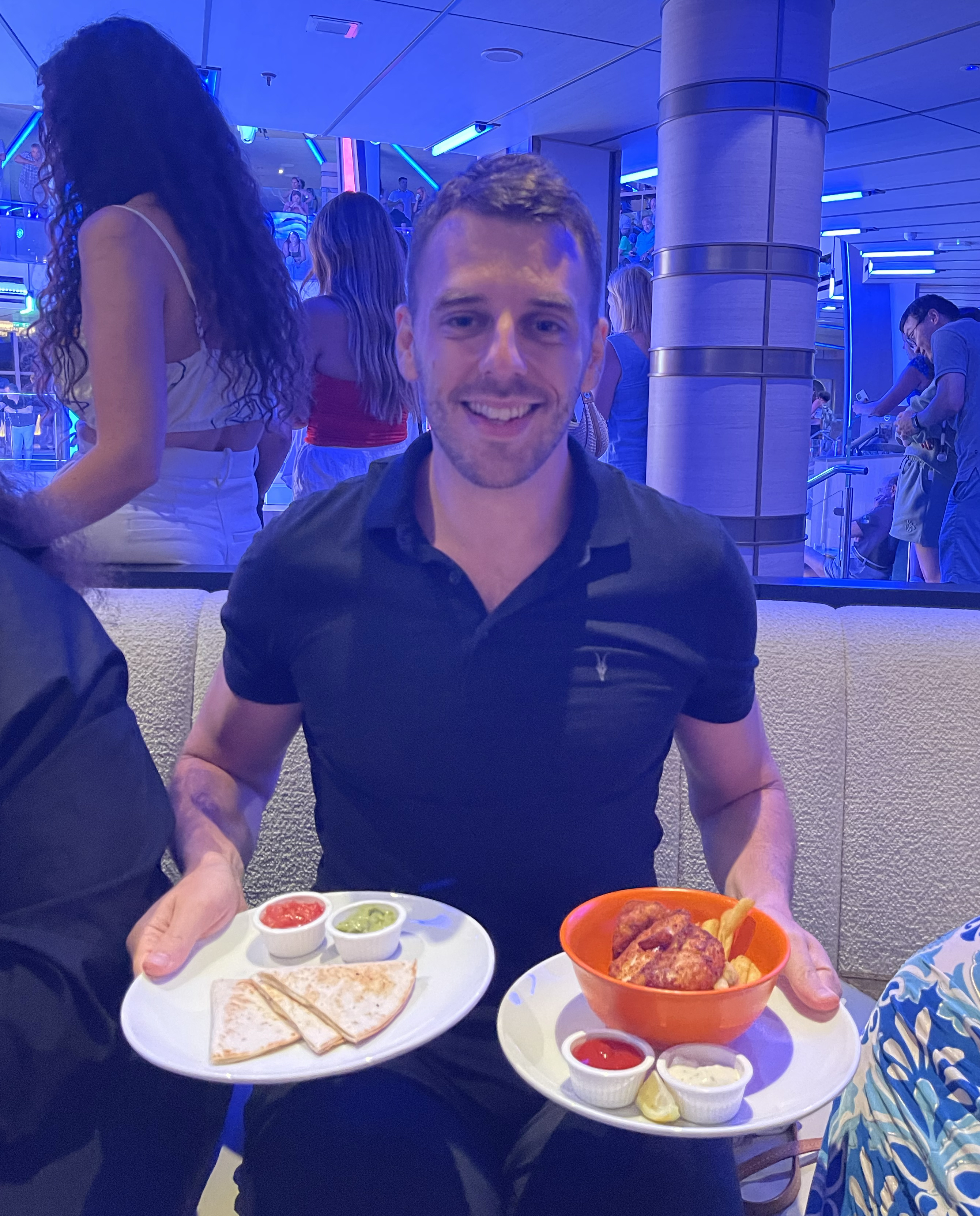 Me holding two plates of food, one with quesadillas and dips, the other with a bowl of chicken wings and dips, sitting in a lively, modern restaurant