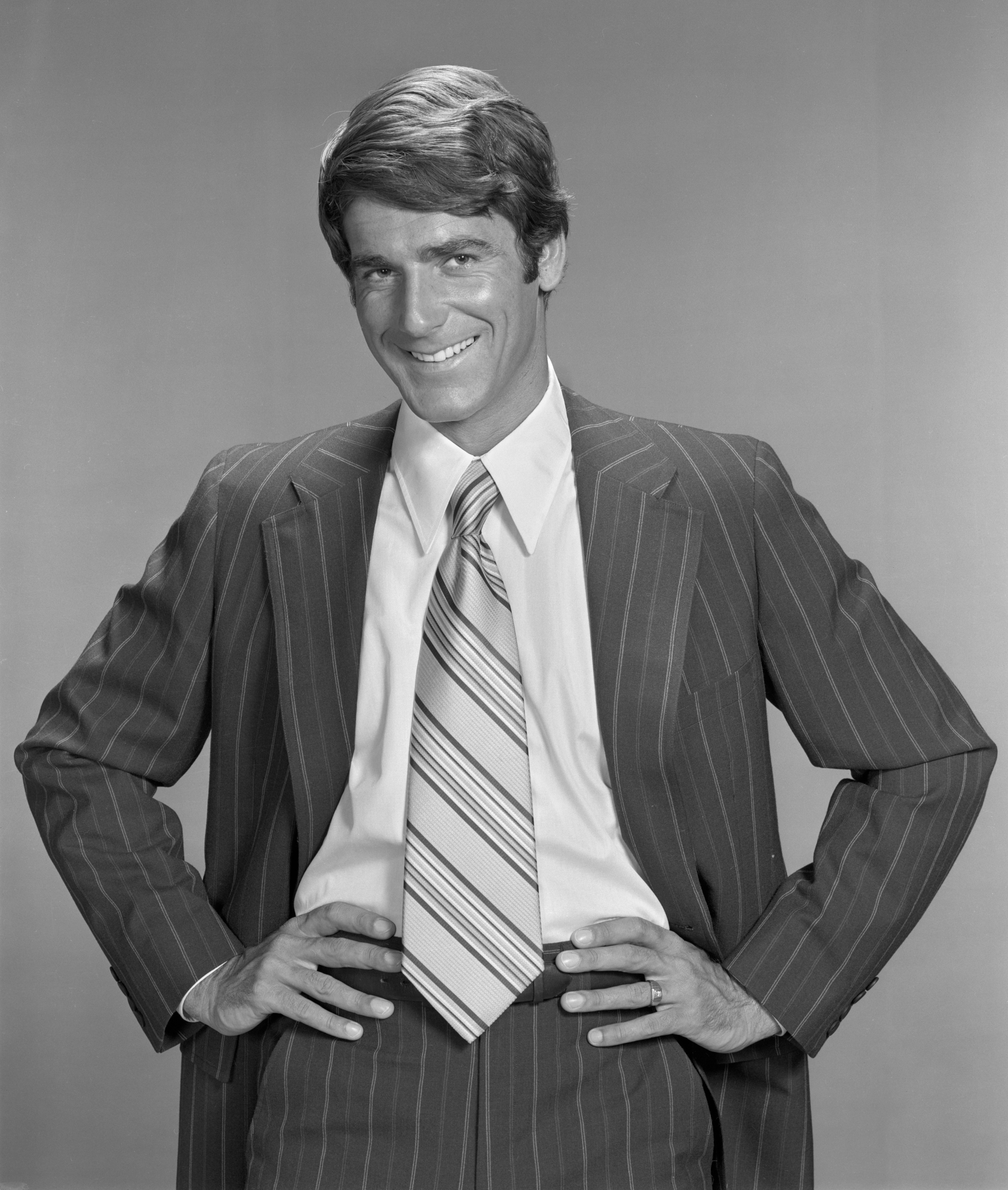 Young Sam Elliott in a pinstripe suit with a striped tie smiling at the camera, with his hands on his hips