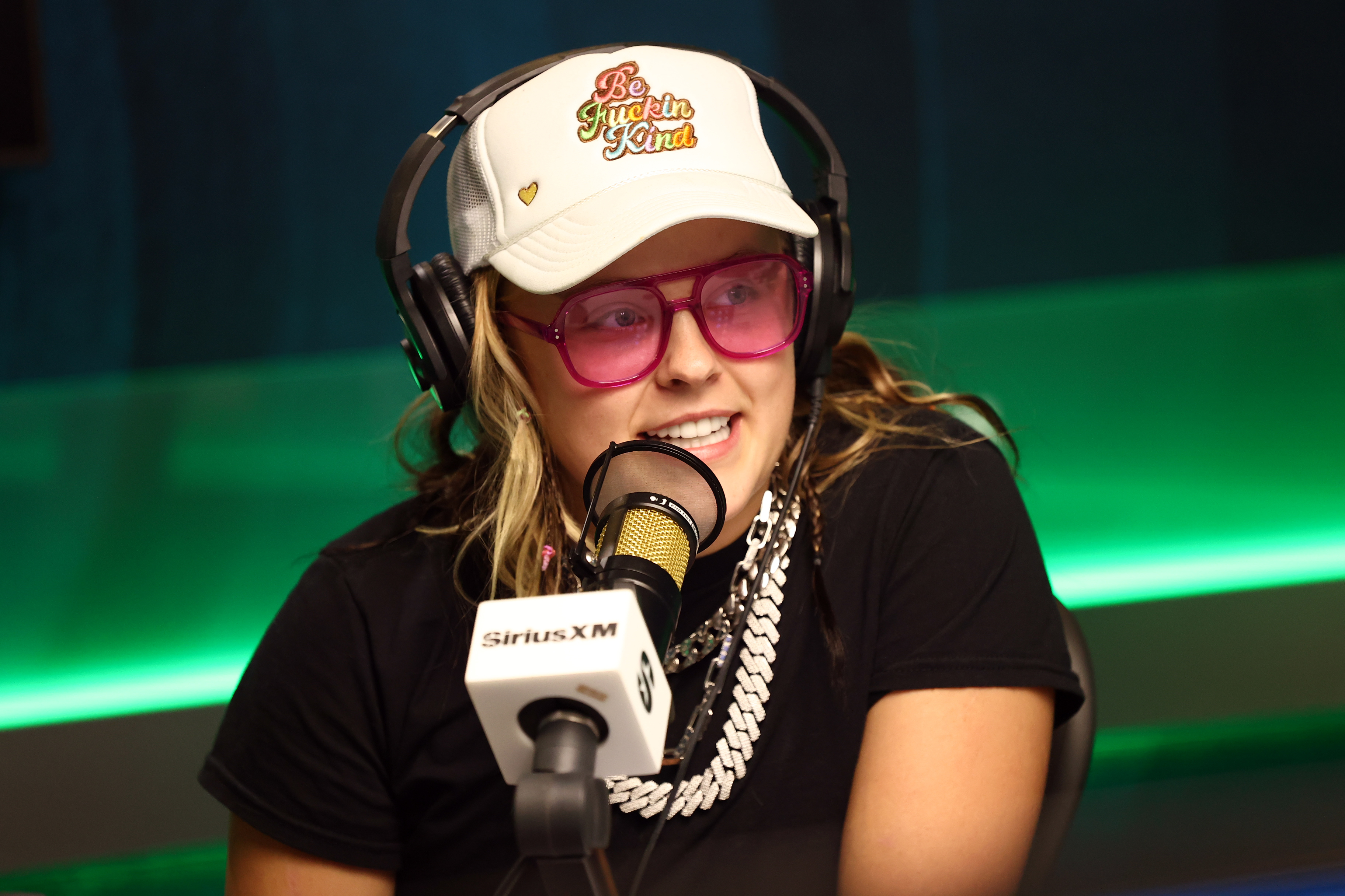 JoJo Siwa speaking on a radio show, wearing a baseball cap, pink glasses, and layered chain necklaces