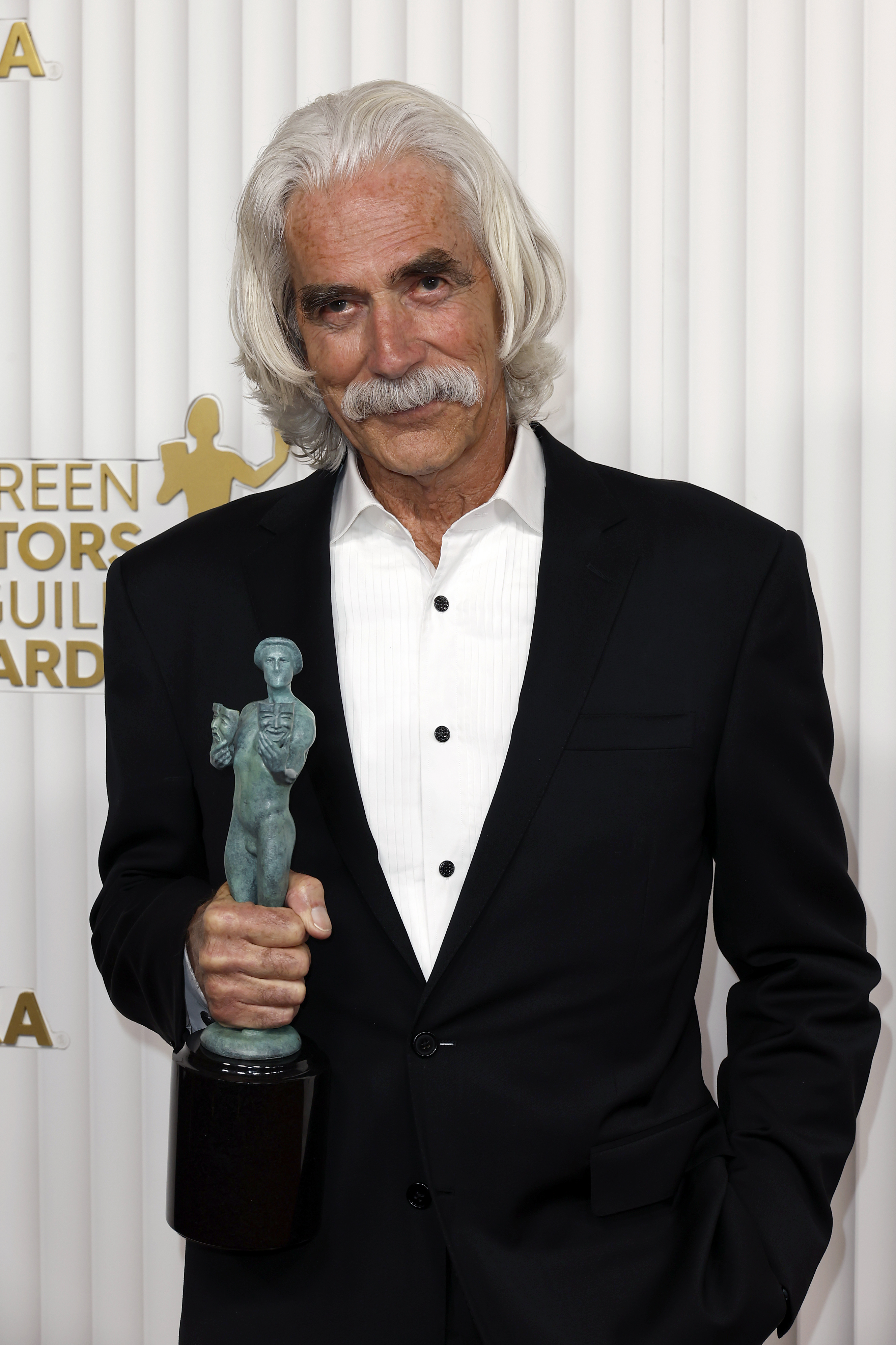 Sam Elliott poses holding an award statue, wearing a dark suit and a white shirt at the Screen Actors Guild Awards