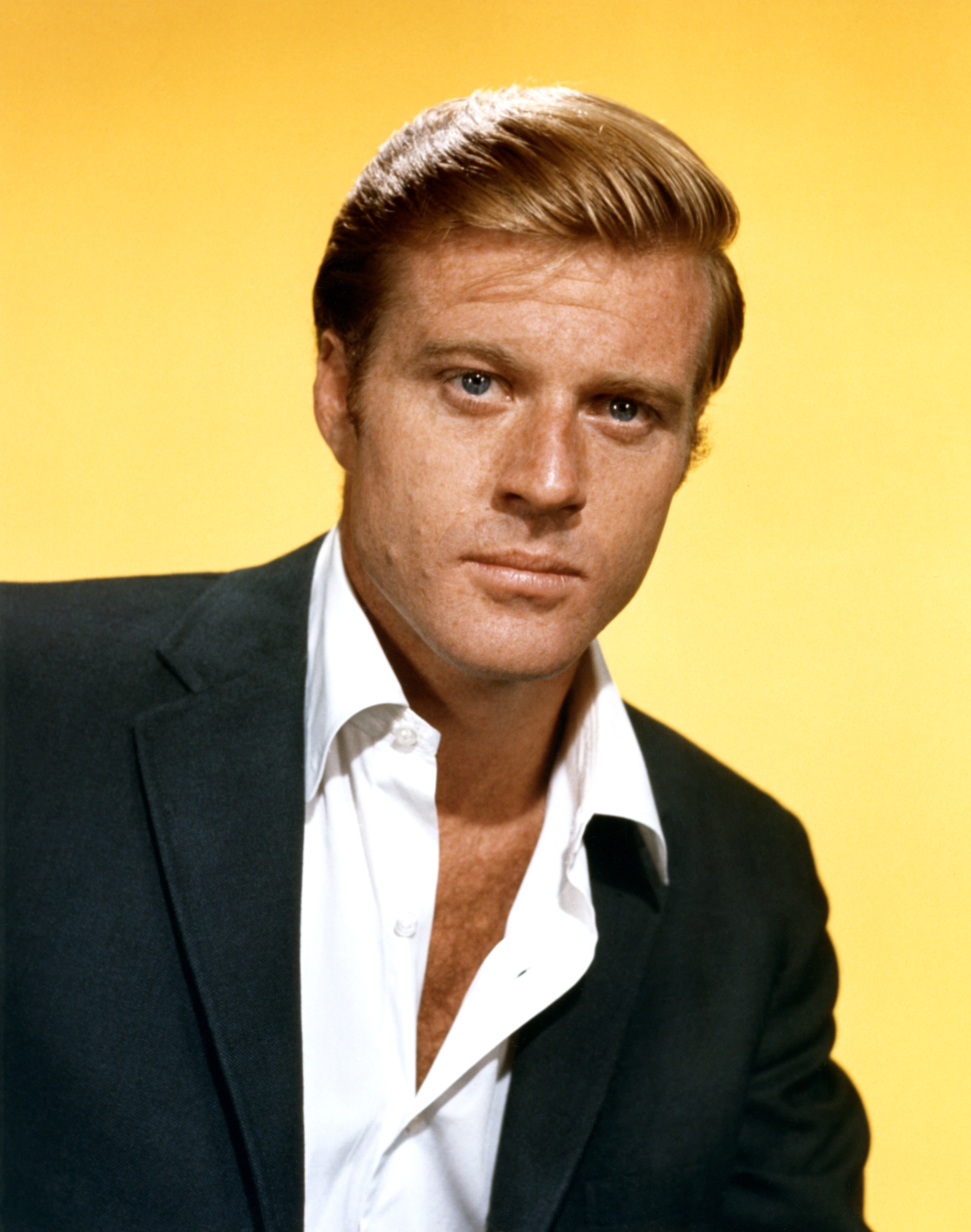 Young Robert Redford in a suit jacket and open-collared shirt, posing against a plain background