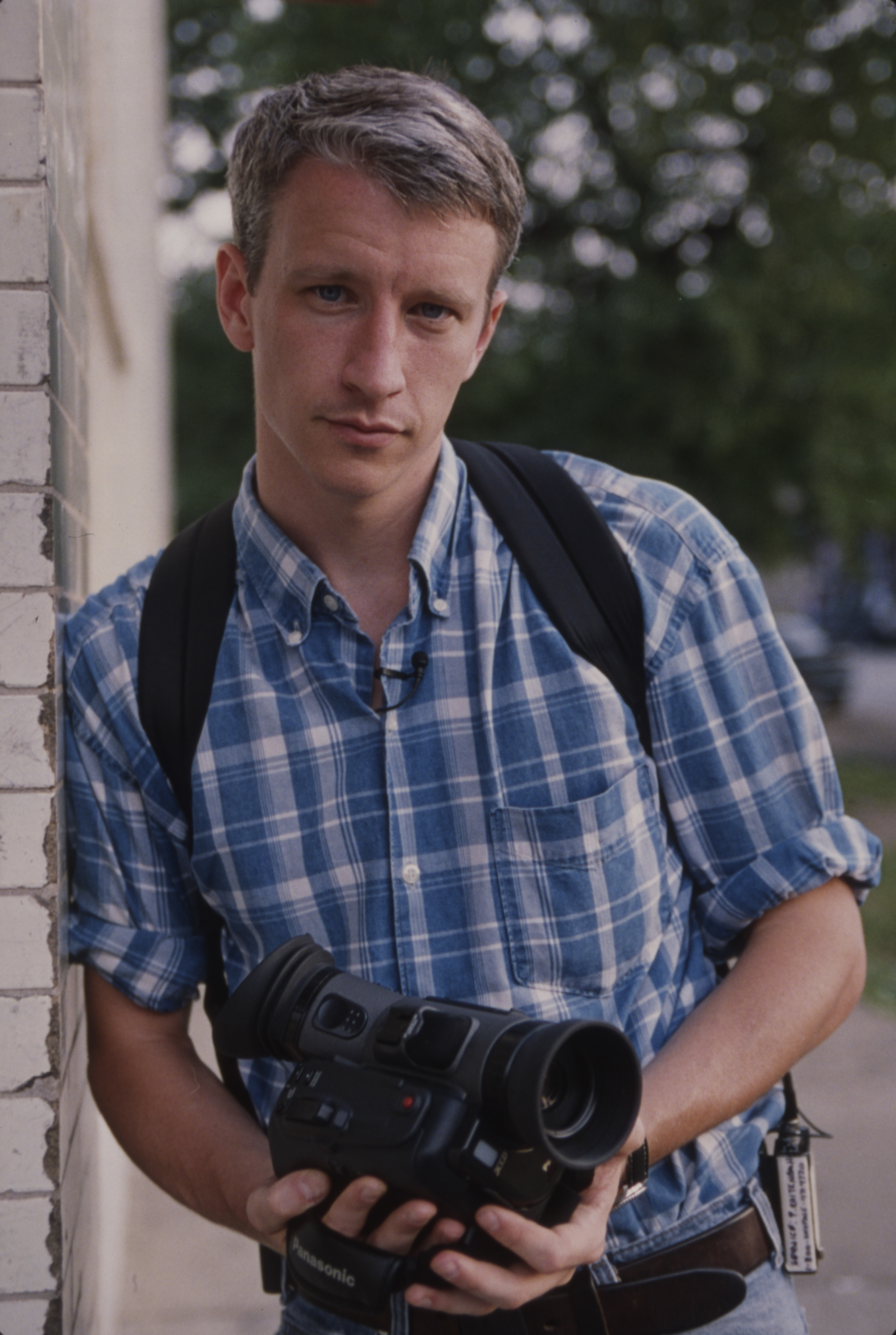 I don't know who this is. A person in a checkered shirt with a backpack holds a video camera, leaning against a brick wall
