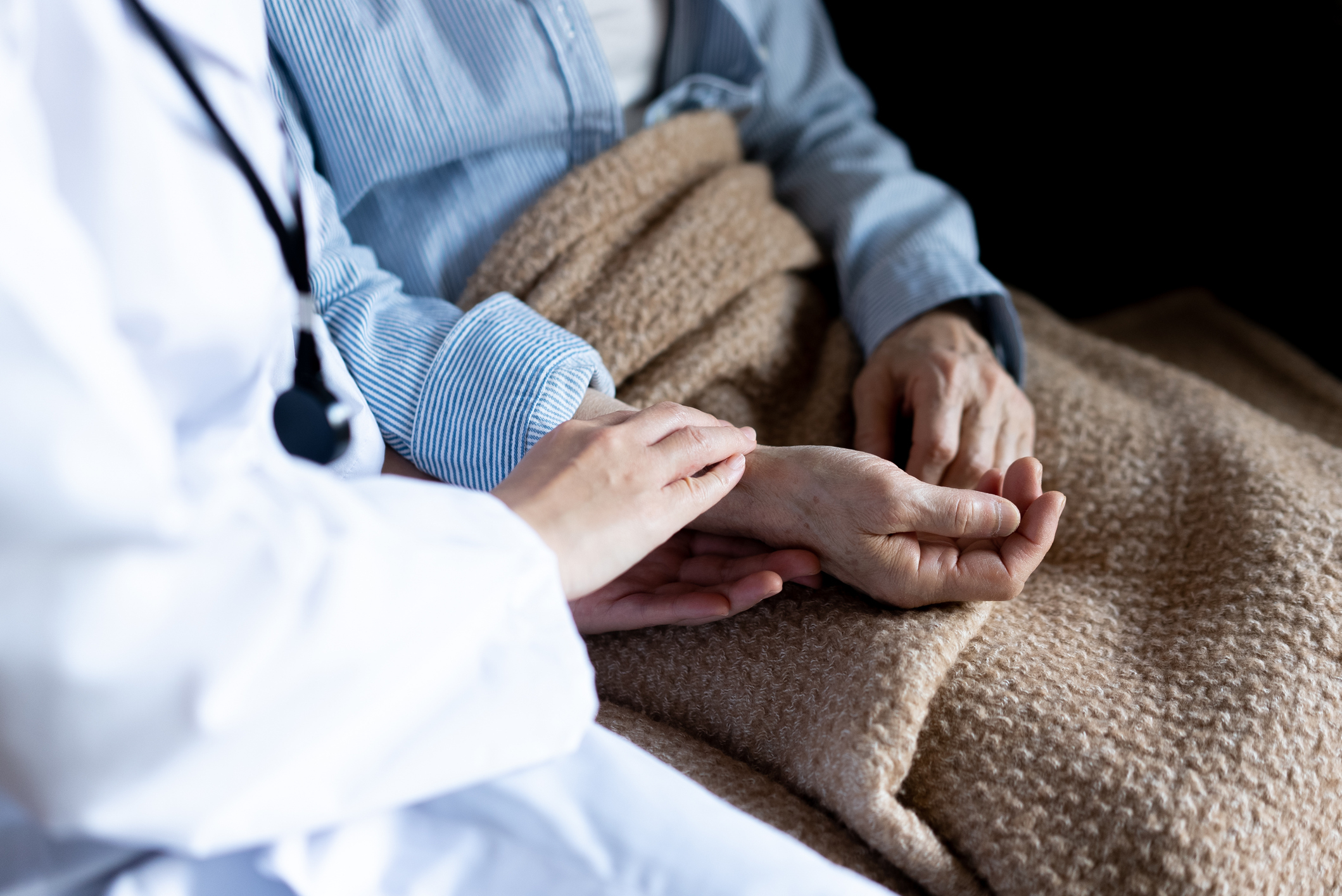 A doctor gently holds the hand of an older patient who is covered with a blanket, showing empathy and care in a clinical setting