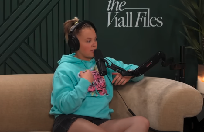 JoJo Siwa sits on a beige couch wearing a hoodie and headphones with a podcast microphone in front of her, on "The Viall Files" podcast