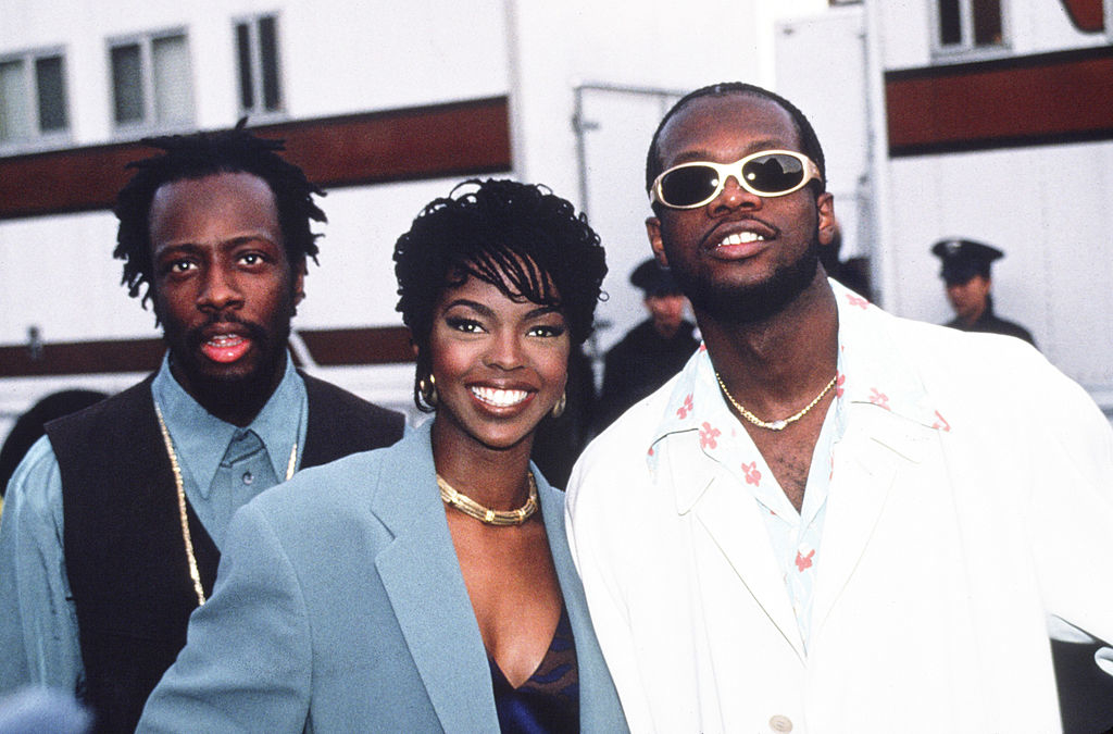 Wyclef Jean, Lauryn Hill, and Pras Michel are dressed stylishly at an event, with Lauryn Hill wearing an elegant blazer and Pras Michel in sunglasses and a white jacket