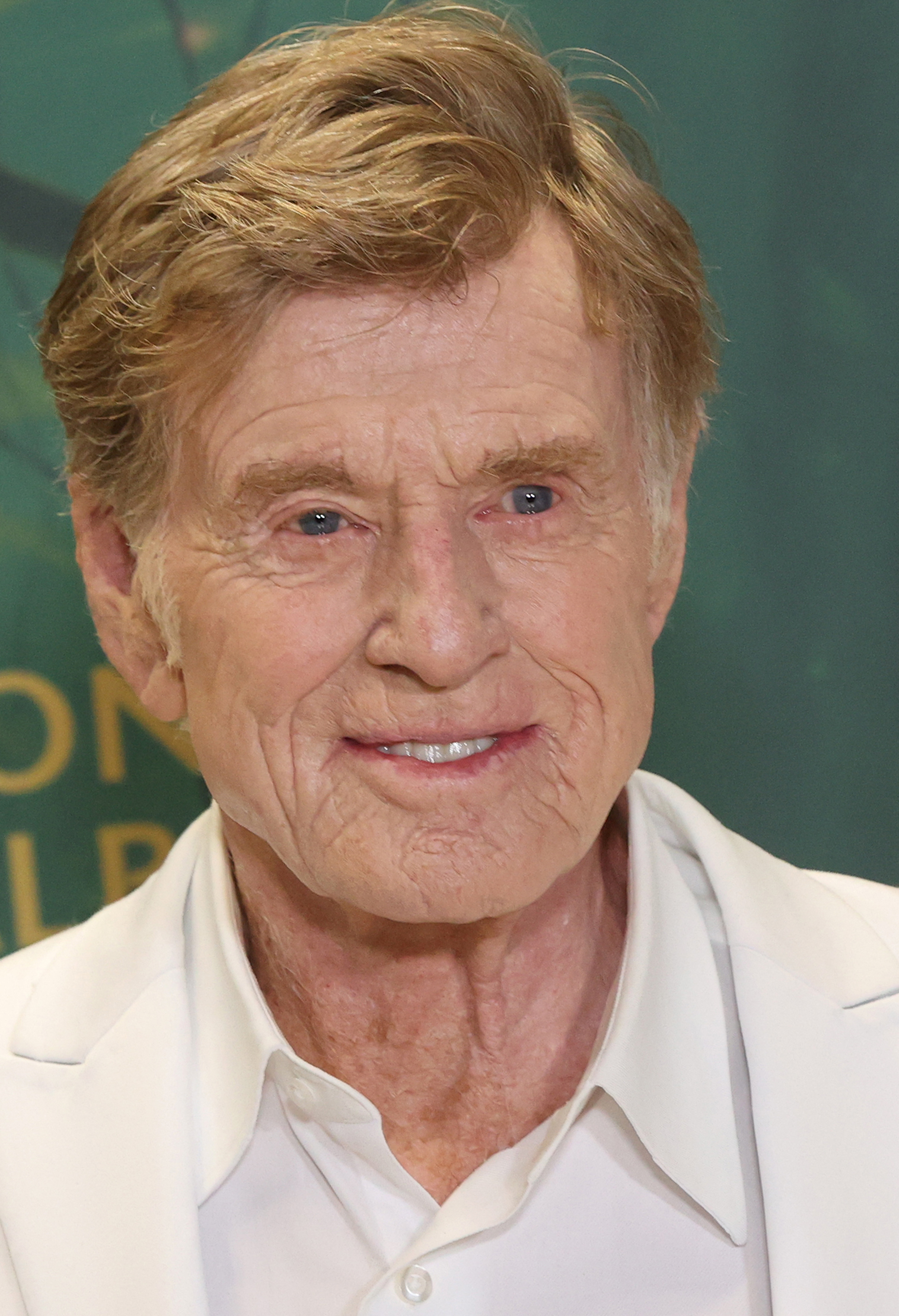 Robert Redford smiles, dressed in a white suit jacket over a white shirt