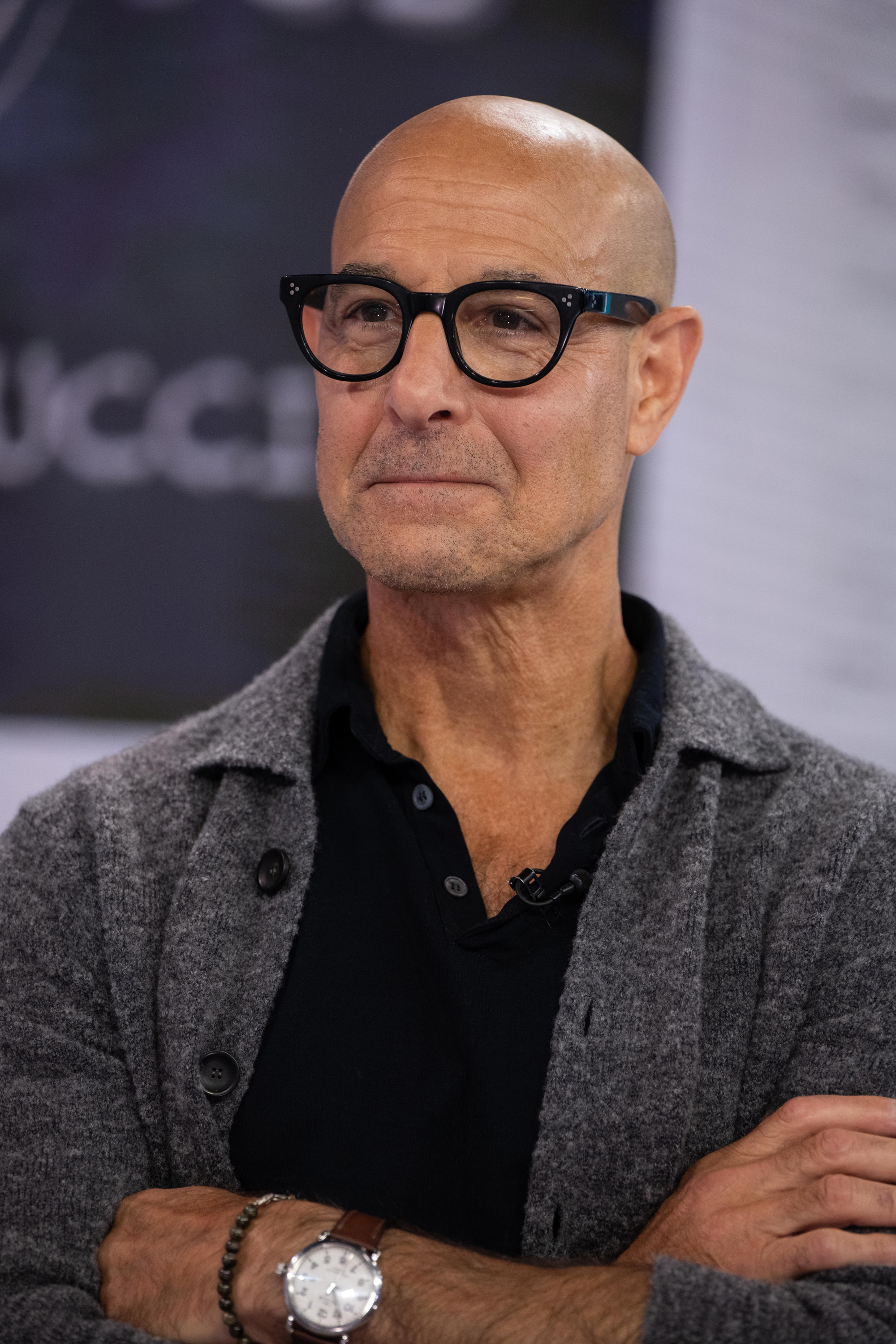 Stanley Tucci, wearing glasses, a black shirt, and a gray cardigan, with arms crossed at a public event
