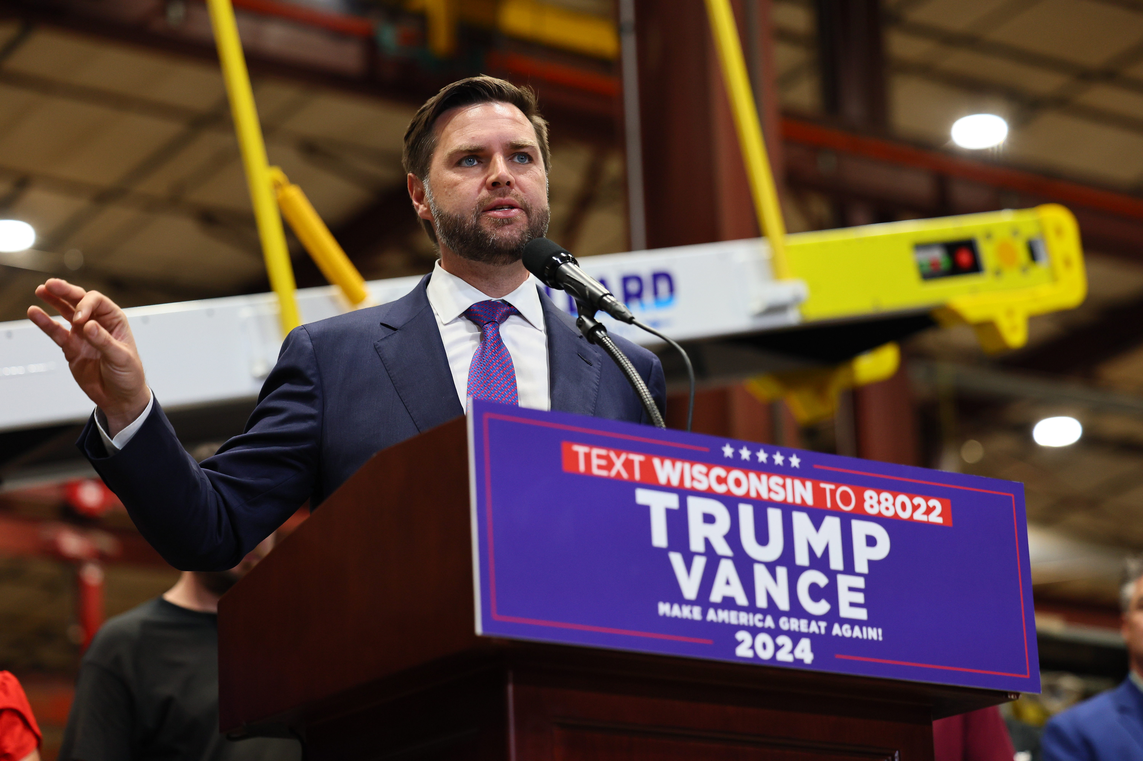 J.D. Vance speaks at a podium with a sign reading &quot;TEXT WISCONSIN TO 88022 TRUMP VANCE 2024.&quot; He is in a suit and tie at an indoor event
