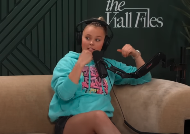 JoJo Siwa in a casual outfit with a hoodie and shorts, speaking into a microphone on The Viall Files podcast