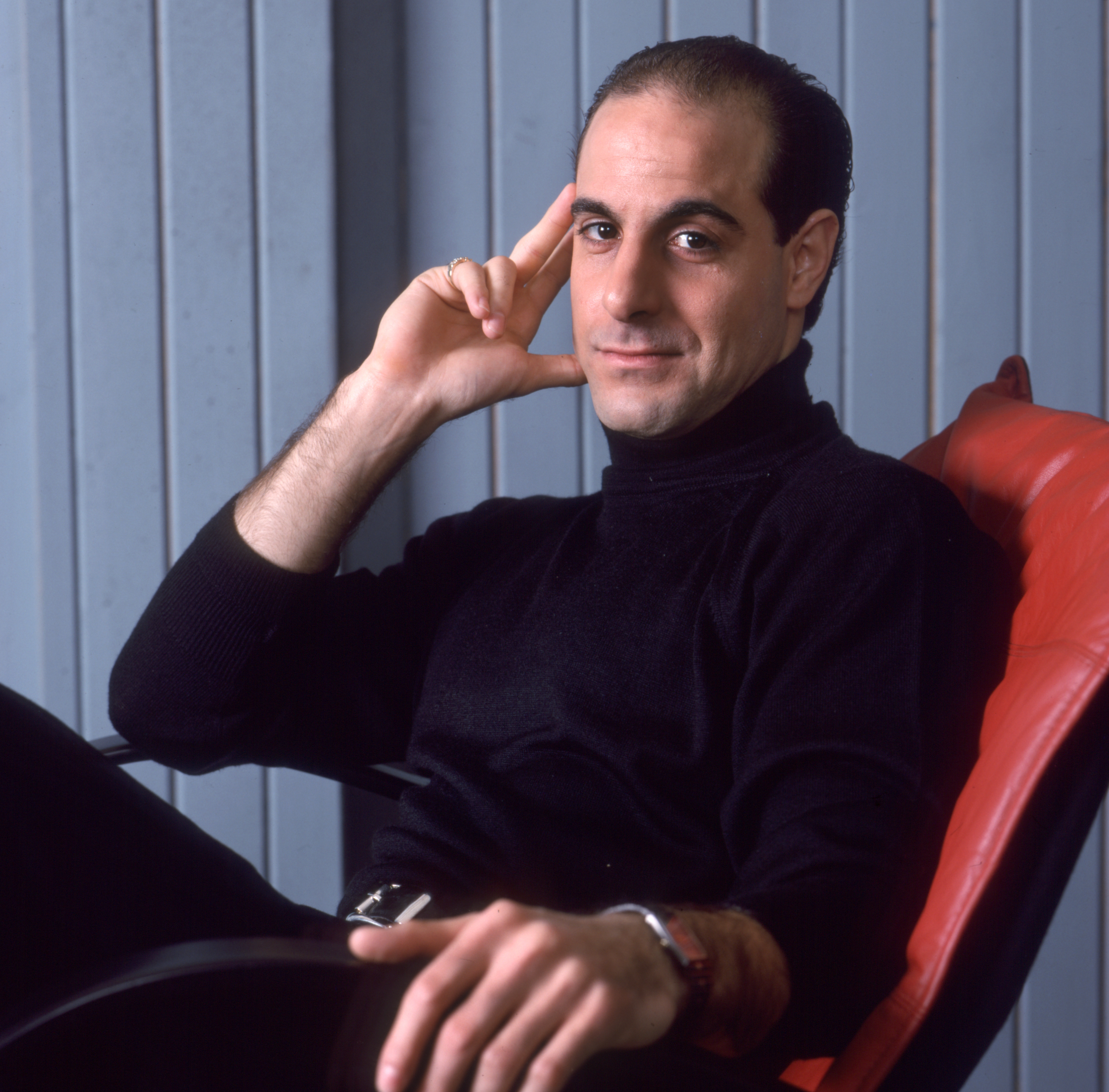 Young Stanley Tucci sits casually in a red chair, wearing a black turtleneck. His left hand is resting on his face and his right hand on his lap