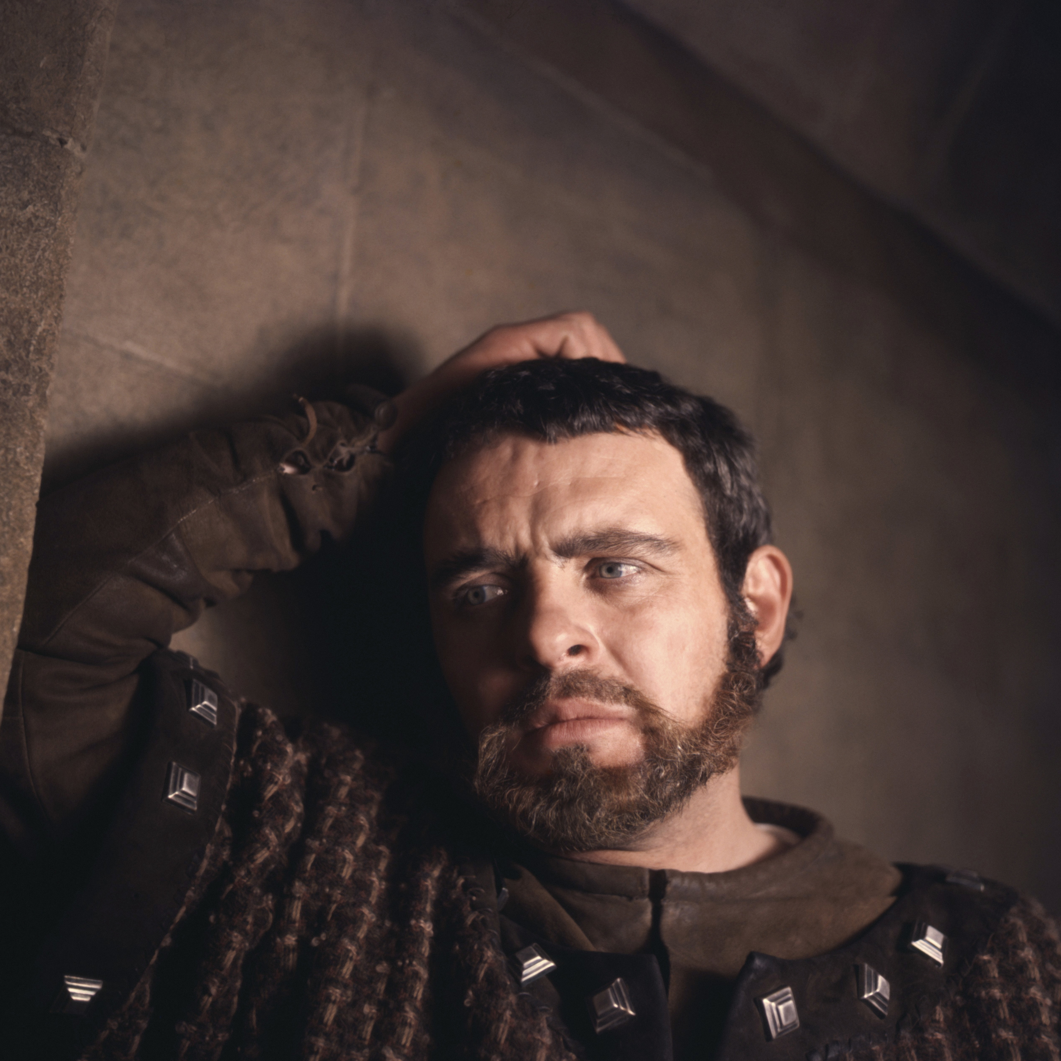 Young Anthony Hopkins with a beard, wearing studded medieval-style clothing, leans against a stone wall looking pensive