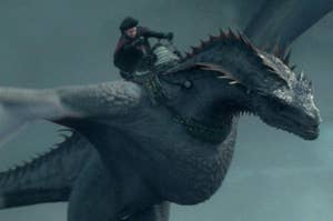 A person rides a large, gray dragon mid-flight. The person is dressed in medieval-style clothing, holding onto the dragon's saddle and reins