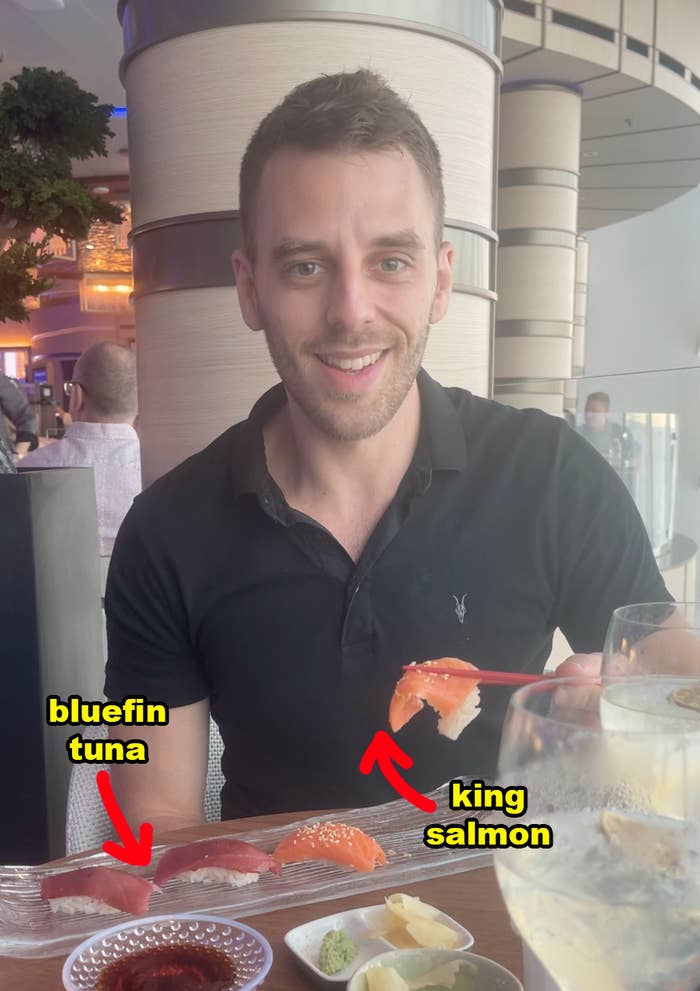 Me smiling while holding a piece of sushi with a restaurant background; sushi on the table is labeled bluefin tuna and king salmon