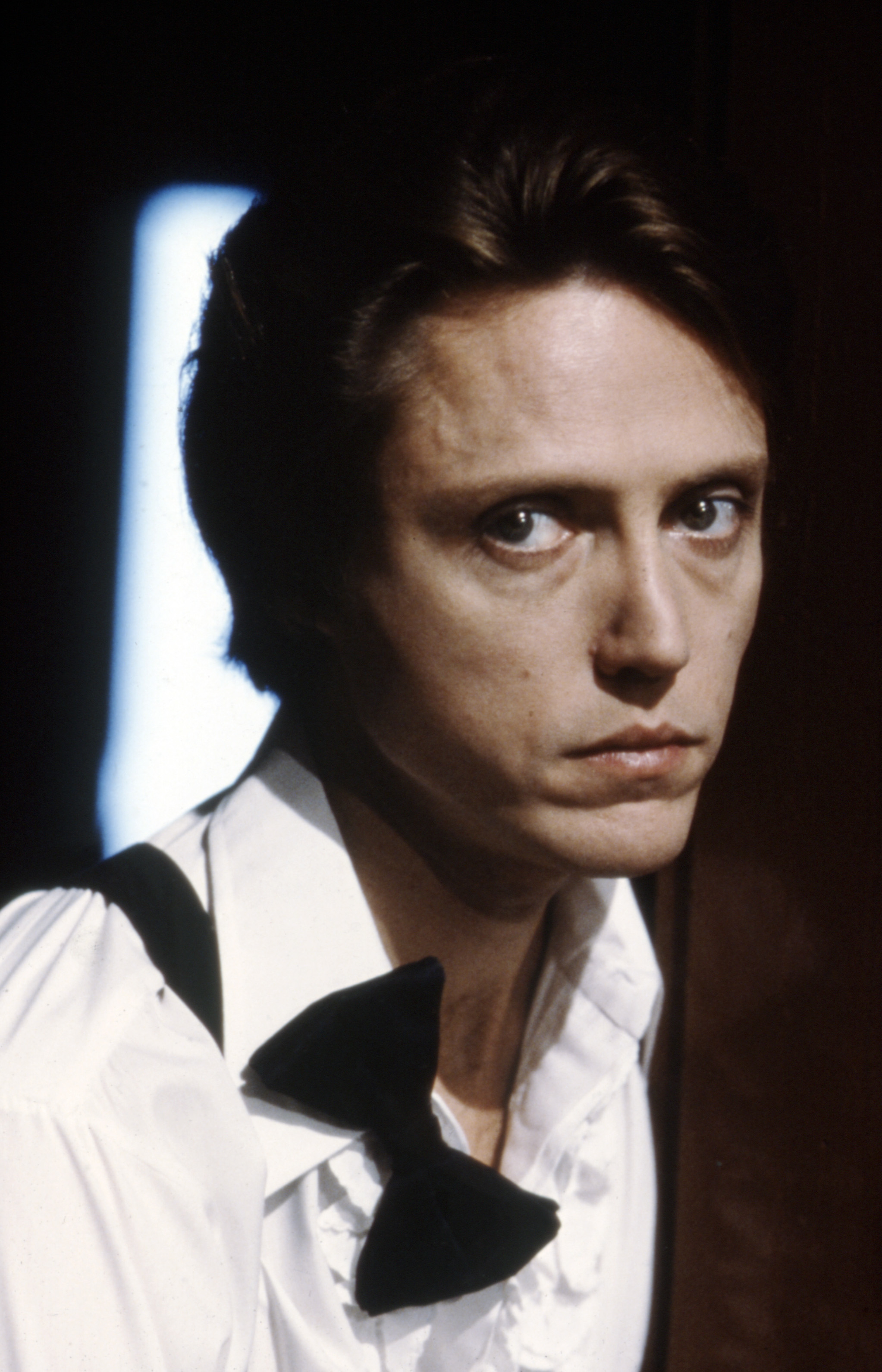 Christopher Walken looks directly at the camera, wearing a white shirt with a black bow tie and suspenders